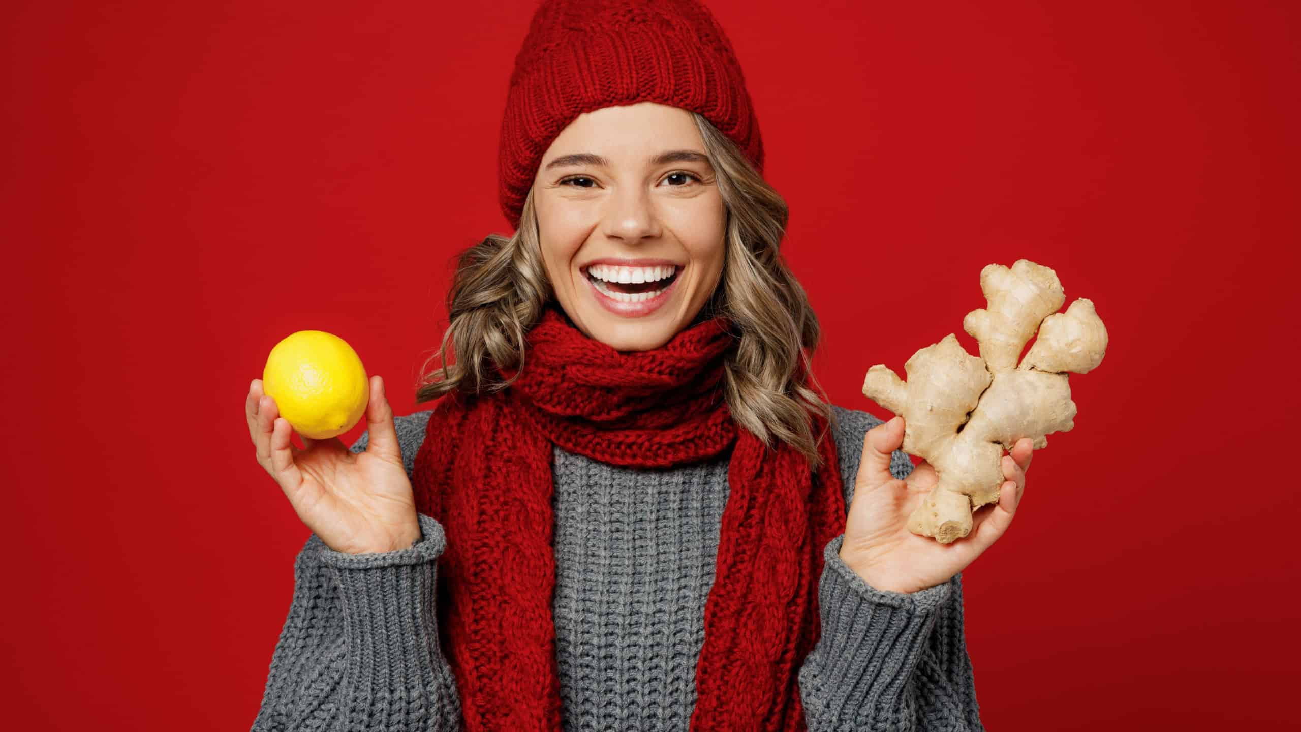 Young smiling happy fun woman wear grey sweater scarf hat hold show lemon ginger root isolated on plain red background studio portrait. Healthy lifestyle ill sick disease treatment cold season concept