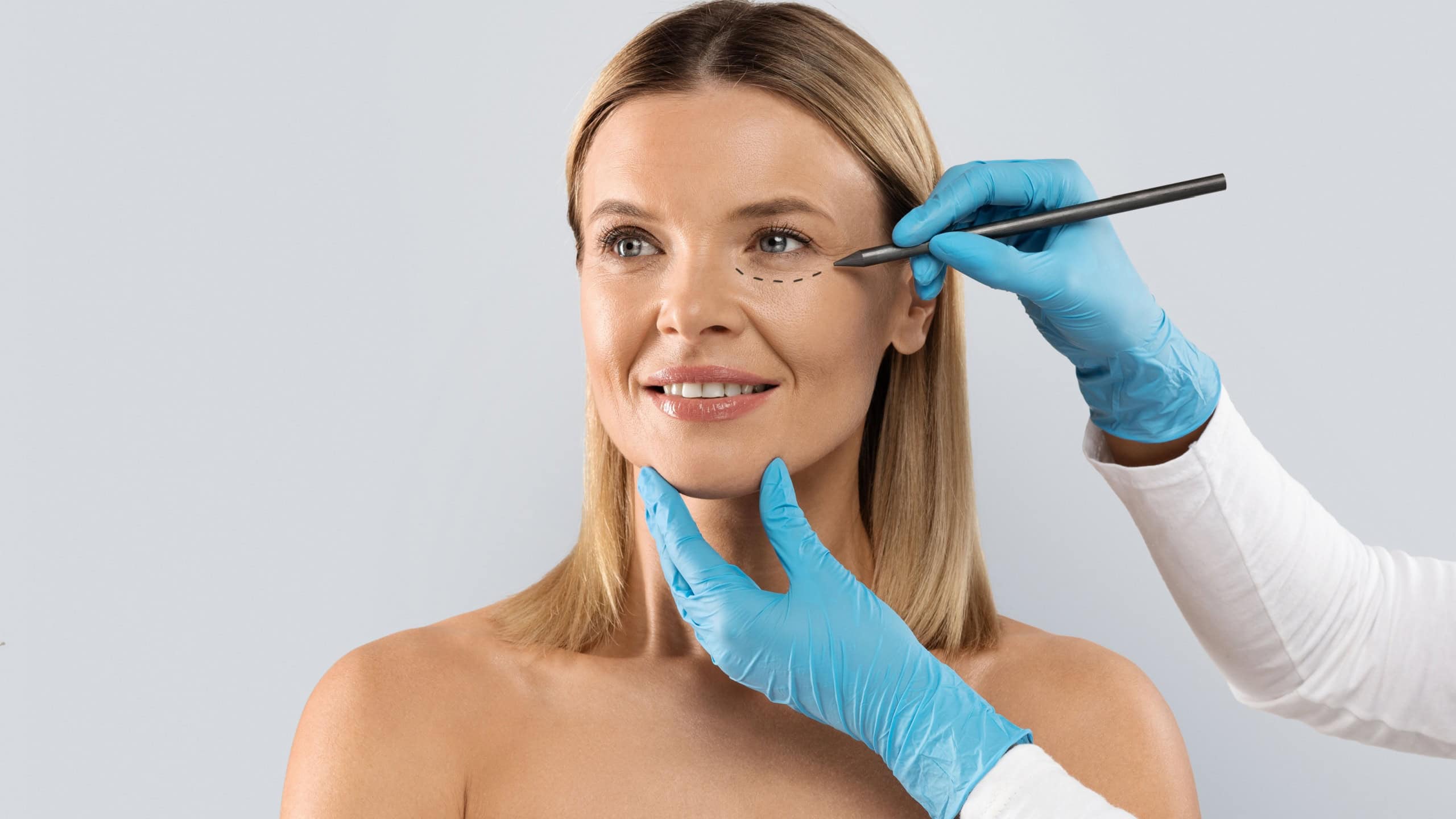 Beautician hands wearing blue medical gloves drawing contours with black pen on woman face before beauty treatment, middle aged blonde lady getting beauty injections, isolated on grey, copy space