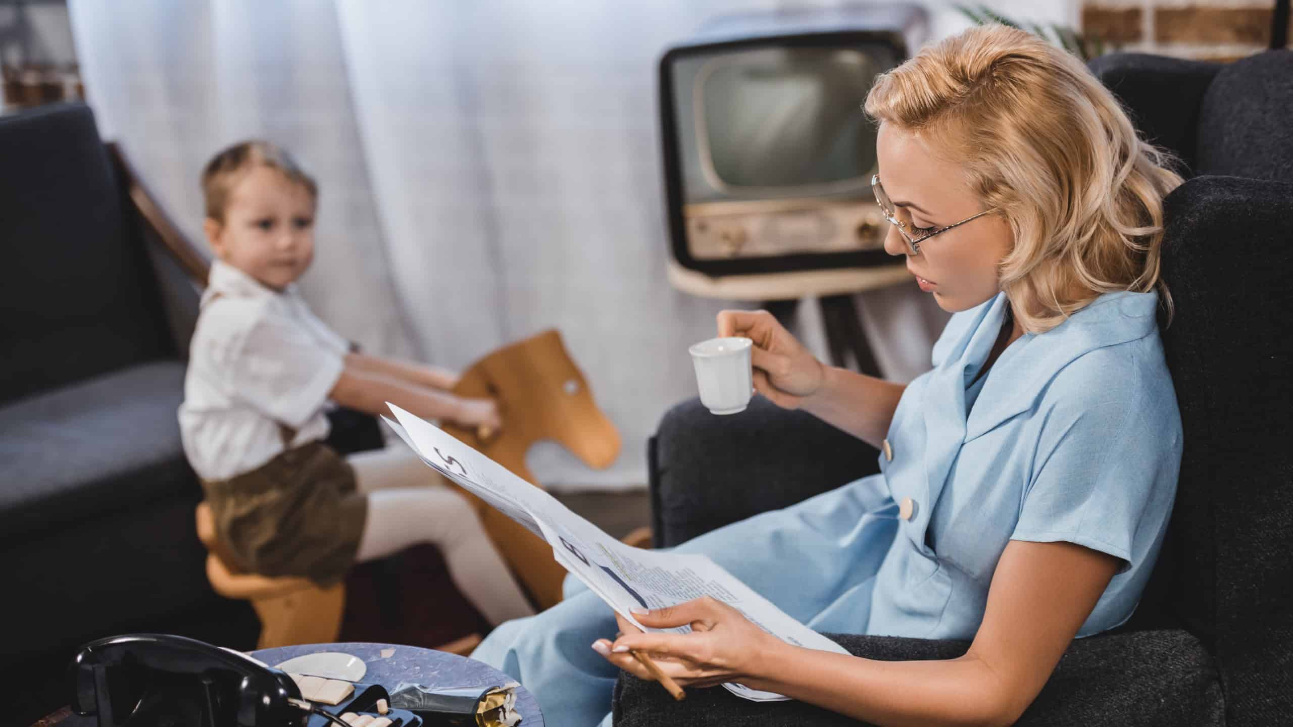 woman in eyeglasses holding cup of coffee and reading newspaper while little son sitting on rocking horse behind, 50s style family