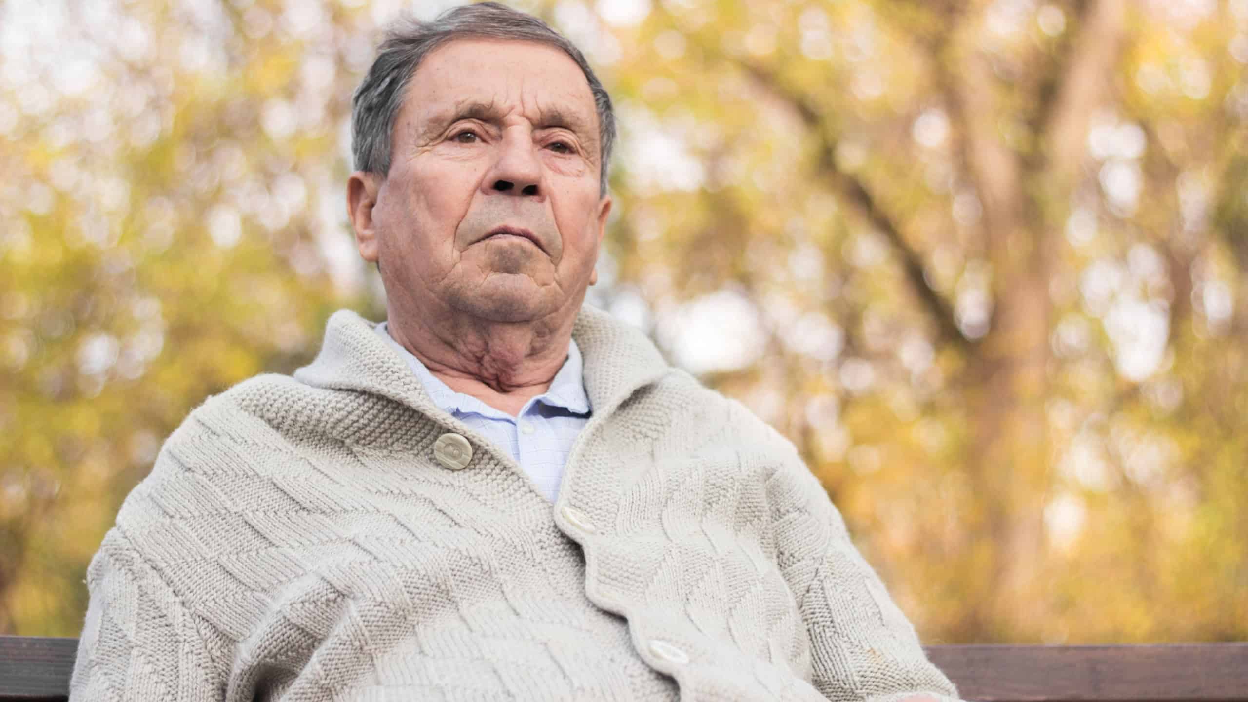 Portrait of a pensive senior man sitting on the bench, in the public park, outdoors. Old man relaxing outdoors and looking away. Portrait of elderly man enjoying retirement