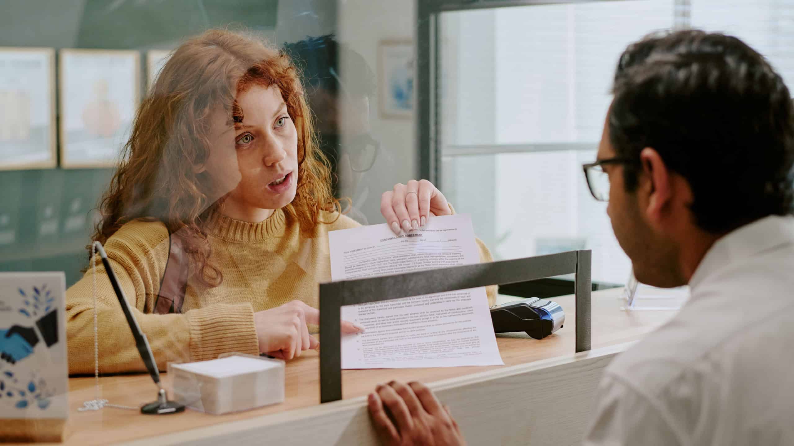 Woman with curly hair discussing financial documents at counter with bank employee focused and engaged in conversation clear glass partition separating them