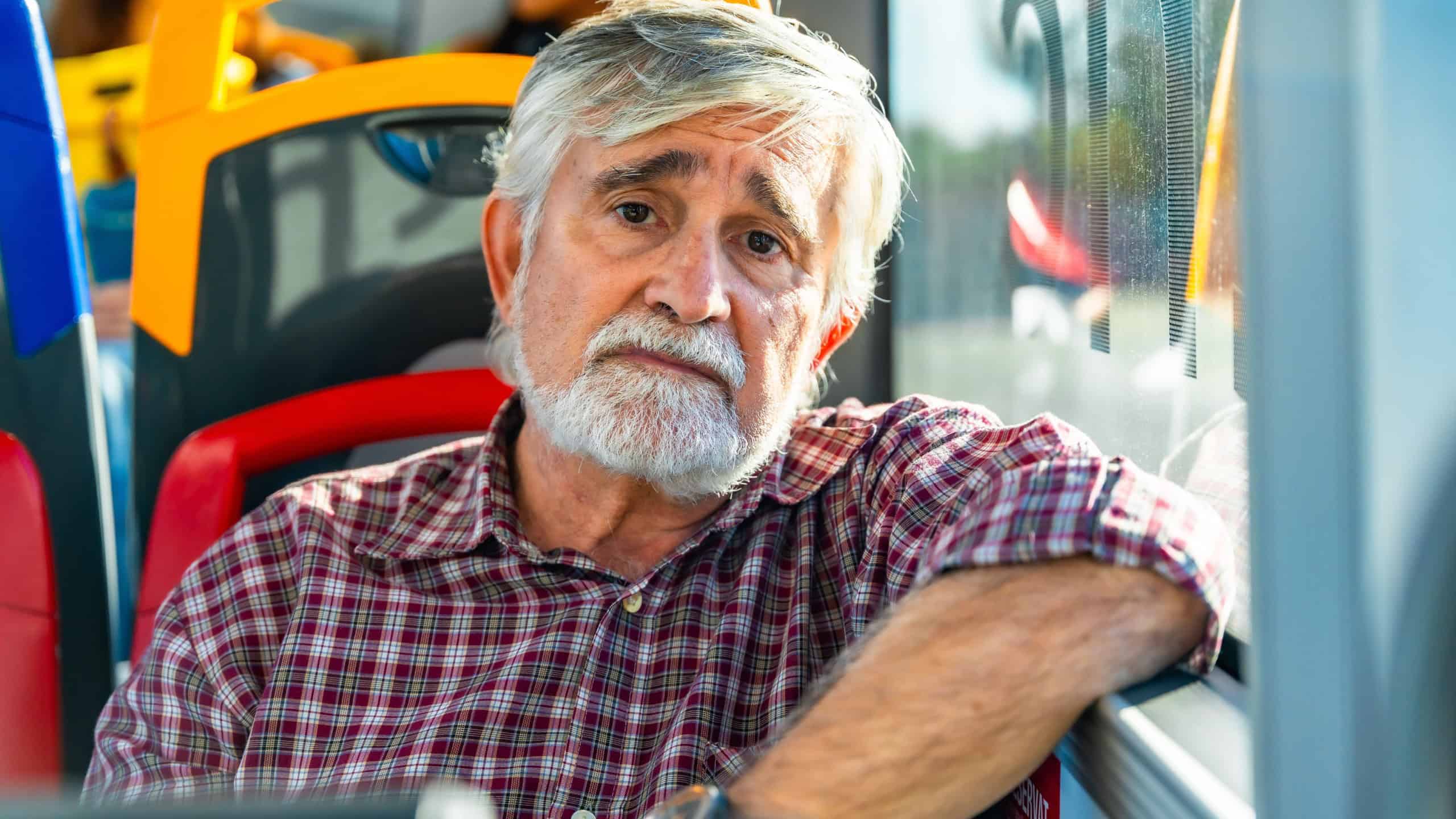 Senior man with white beard and plaid shirt sits by a bus window, gazing pensively at the camera during an urban commute, conveying quiet reflection and daily routine
