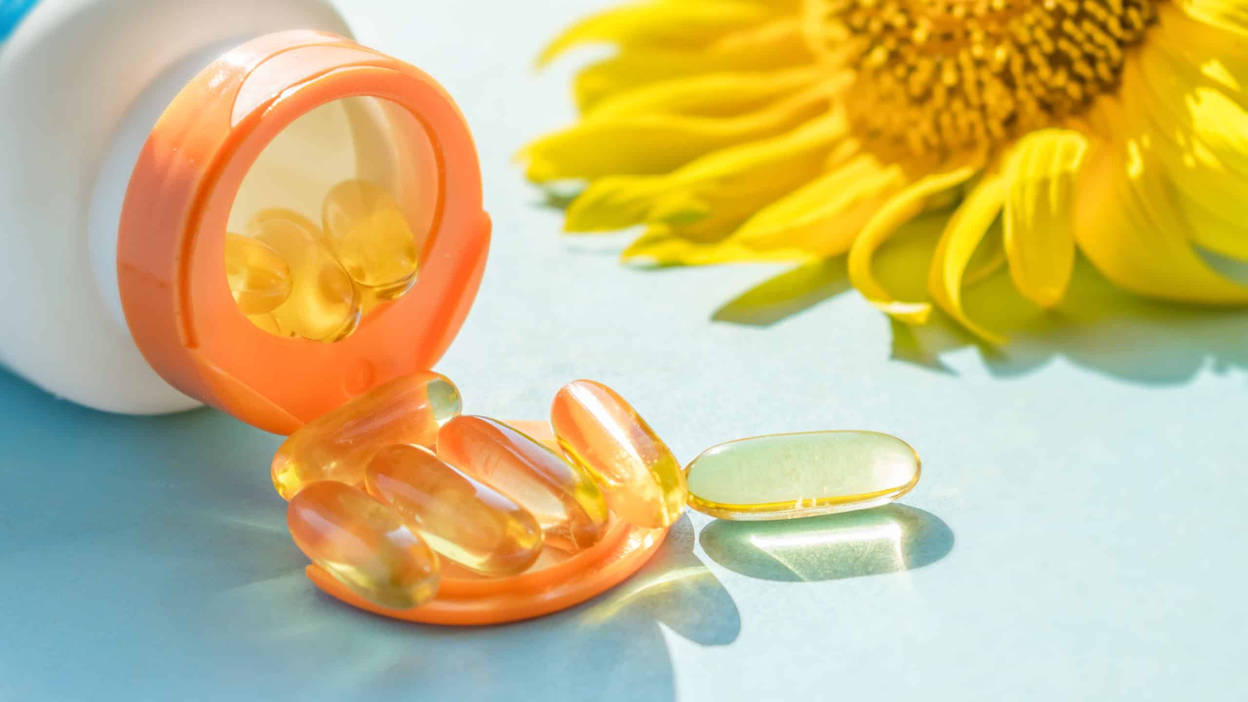 Golden Sunscreen pills with bottle and sunflower on blue background in sunlight. Dietary supplements, protect skin from the sun. Take pill and avoid the burn