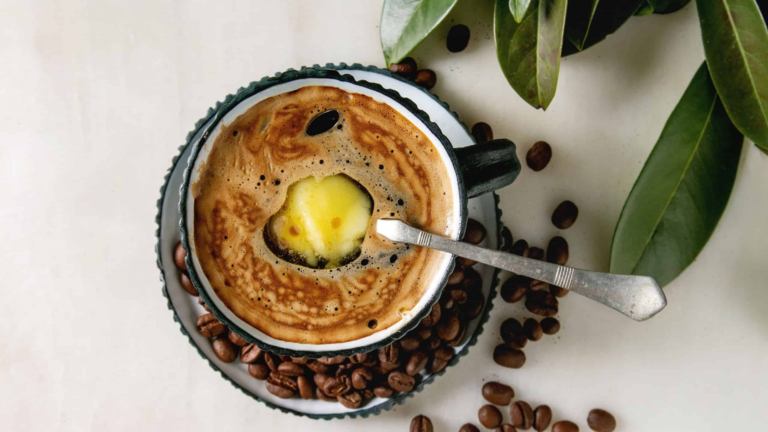 Bulletproof coffee. Keto diet coffee in blue ceramic cup with organic ghee butter in spoon inside with beans and green branch over white marble background. Flat lay, space