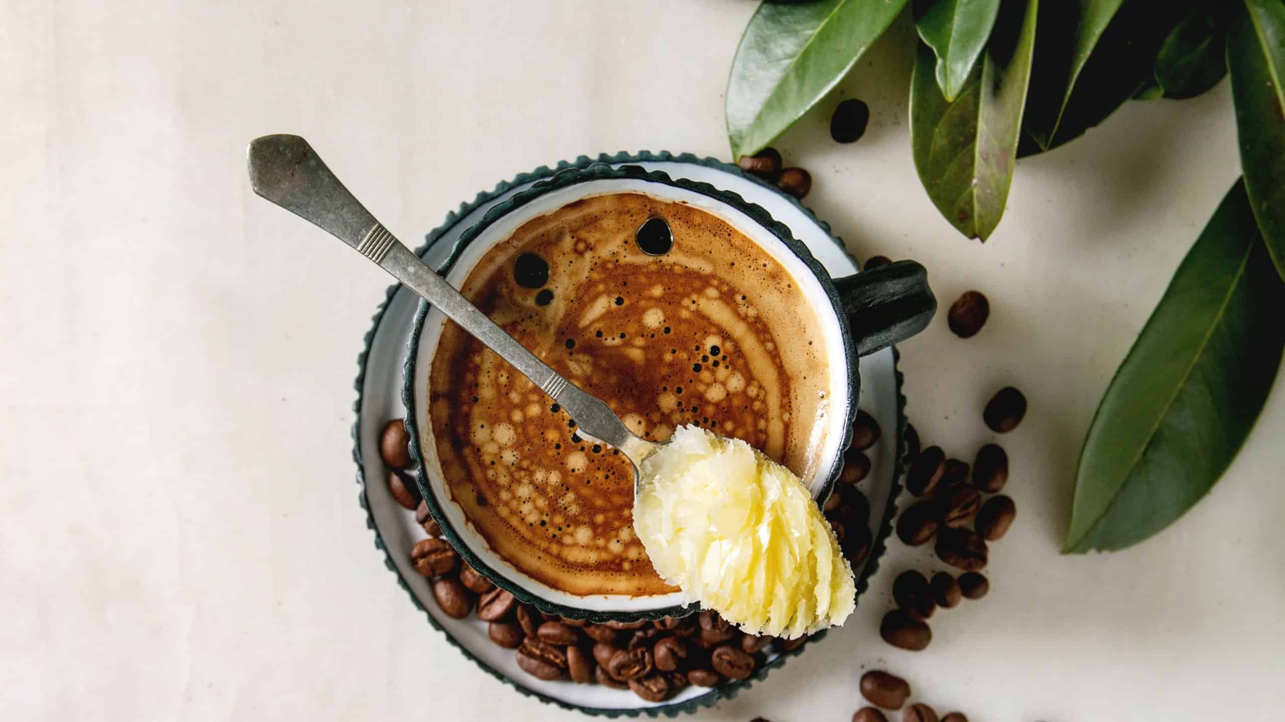 Bulletproof coffee. Keto diet coffee in blue ceramic cup with organic ghee butter in spoon with beans and green branch over white marble background. Flat lay, space