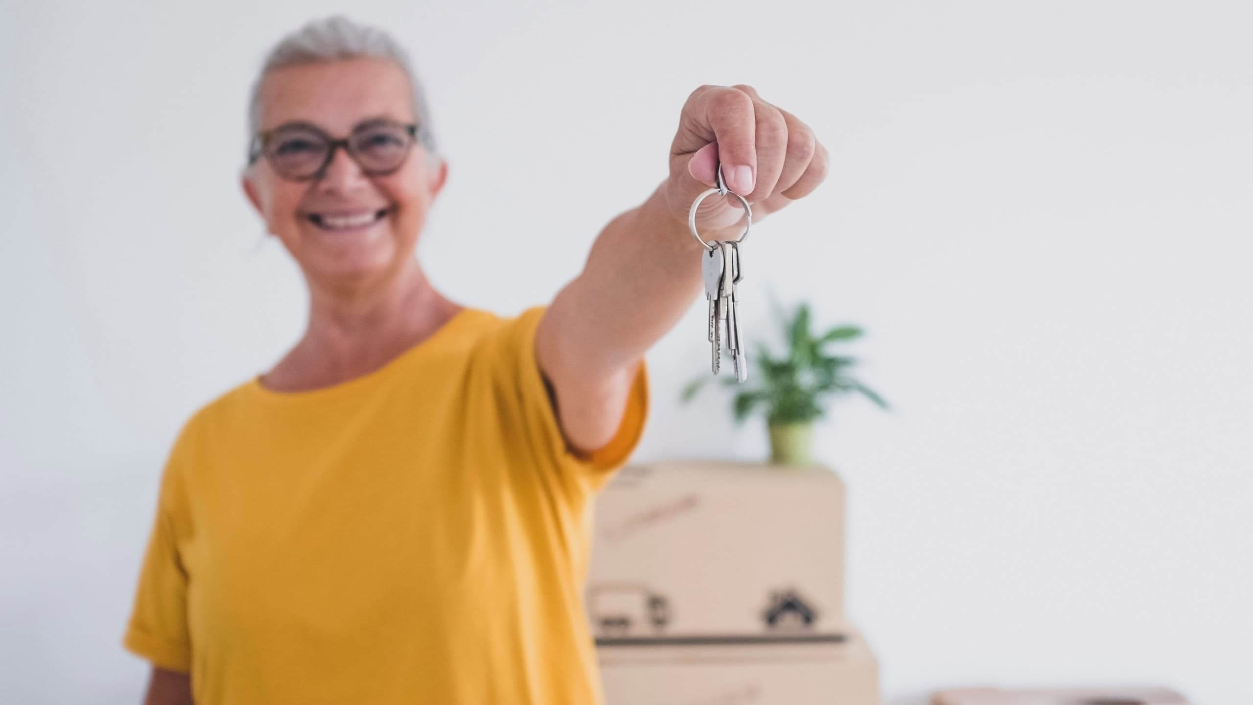 Defocused senior happy woman holding keys of the new apartment standing in the empty home with moving boxes on the floor - concept of active elderly retired enjoying new life