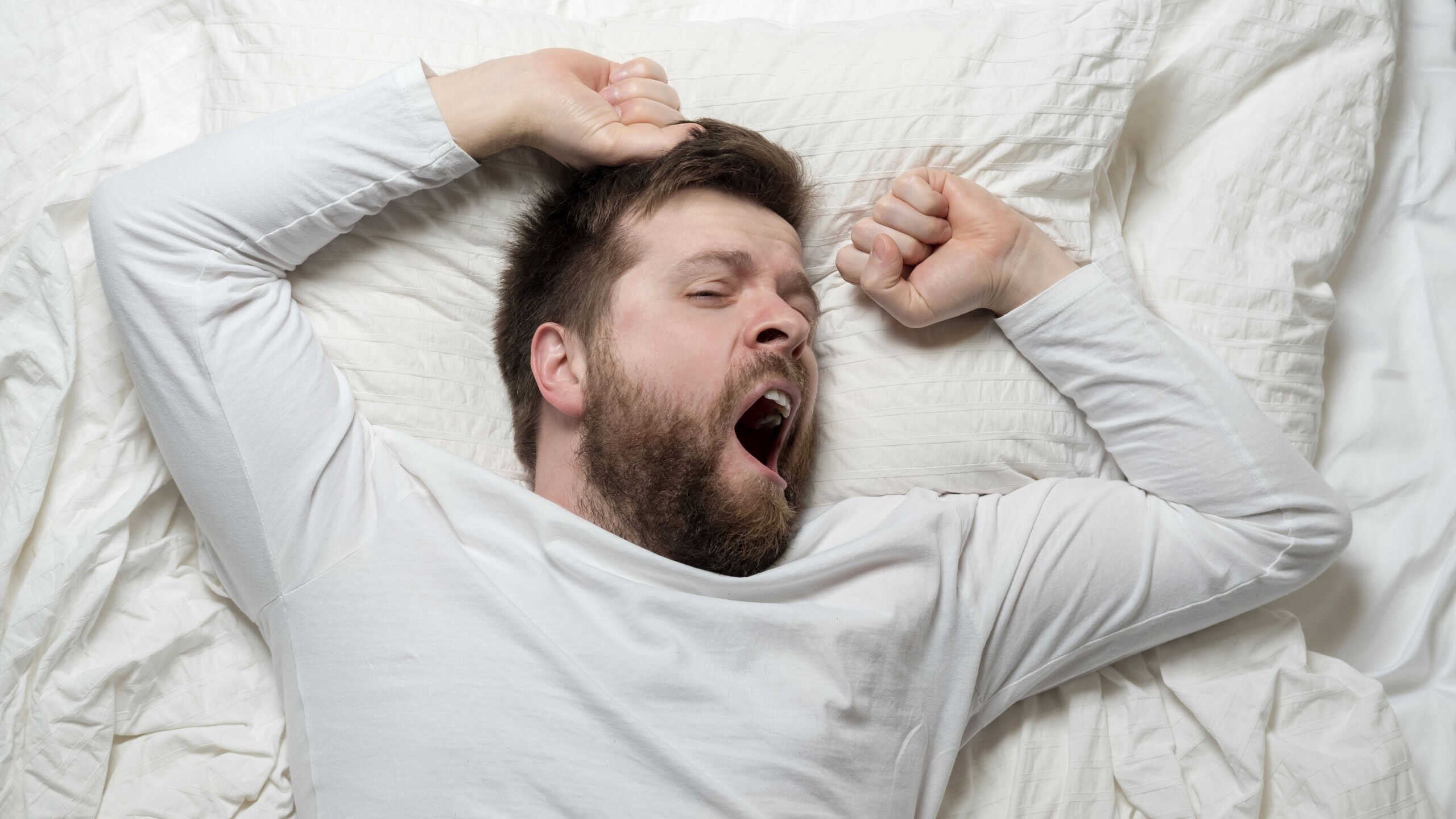 Cute bearded man wakes up from sleep, he yawns and stretches as he lies in bed. Top view. Close-up.