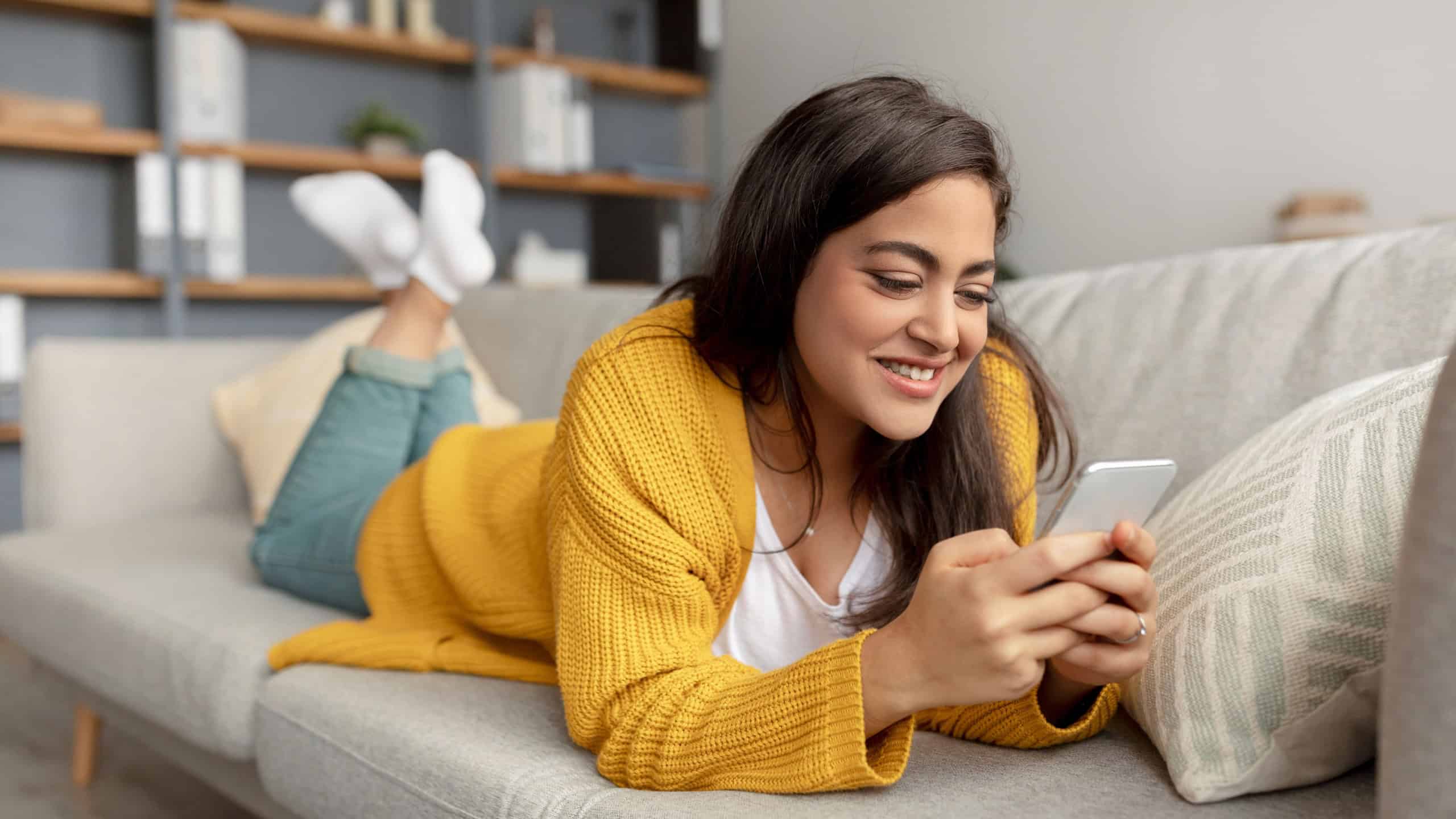 Charming arab lady lying on sofa with smartphone, checking social media, chatting online at home, free space. Happy woman posting on web, watching video on mobile device
