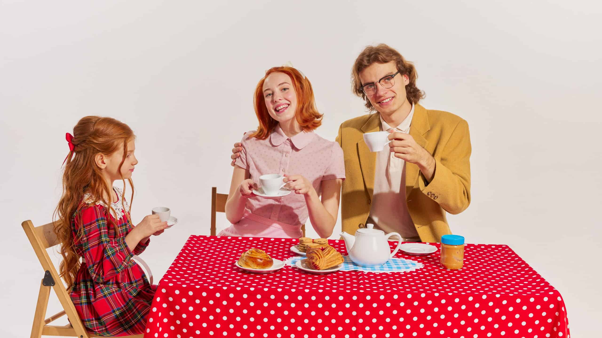 Portrait of happy young family, man, woman and little girl sitting at the table and eating isolated over grey background. Concept of beauty, retro style, fashion, 60s, 70s, family. Copy space for ad