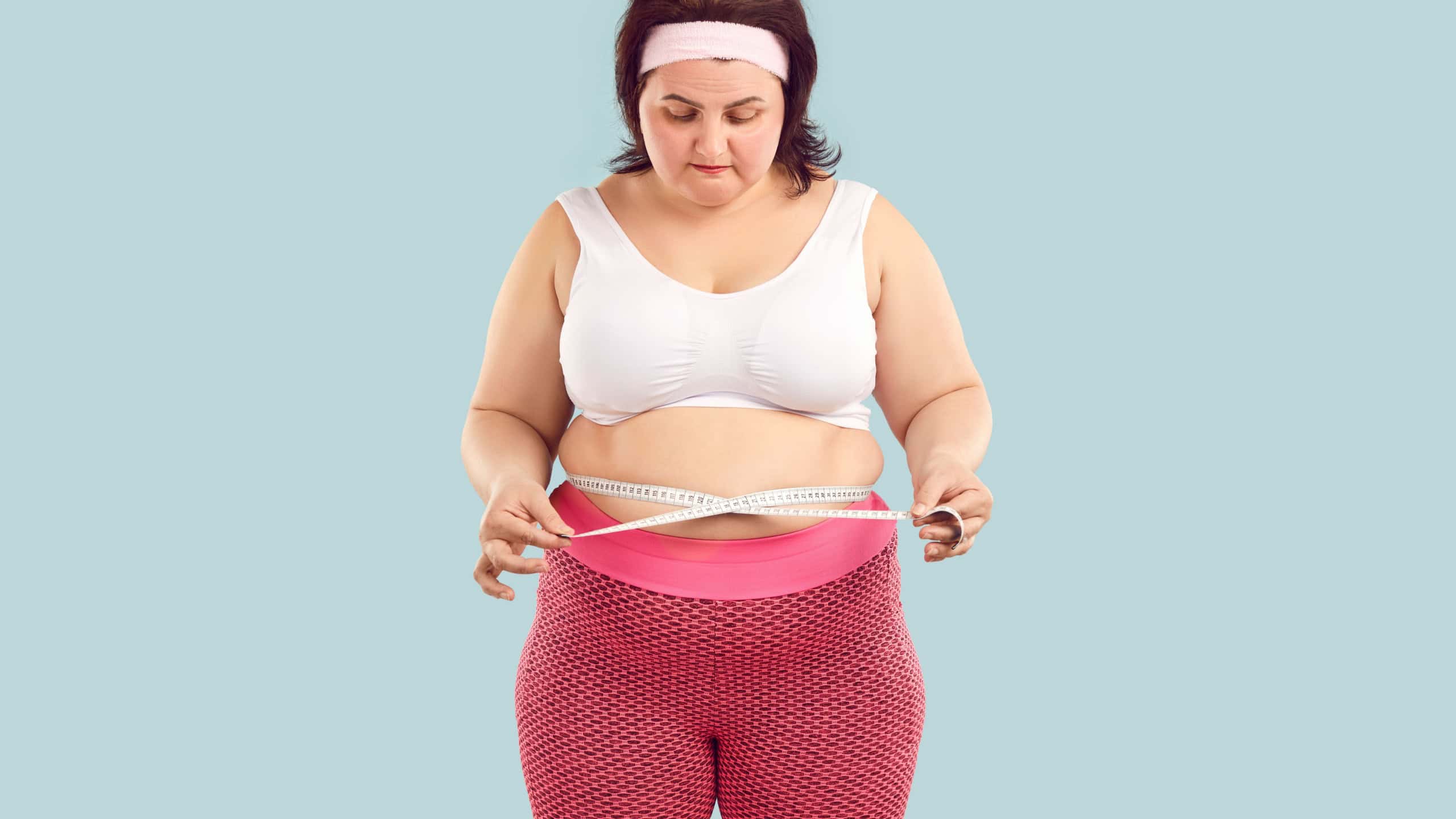 Portrait of a young fat overweight woman wearing sportswear measure the waist with measuring tape isolated on studio blue background. Obesity, weight control, sport and fitness concept.