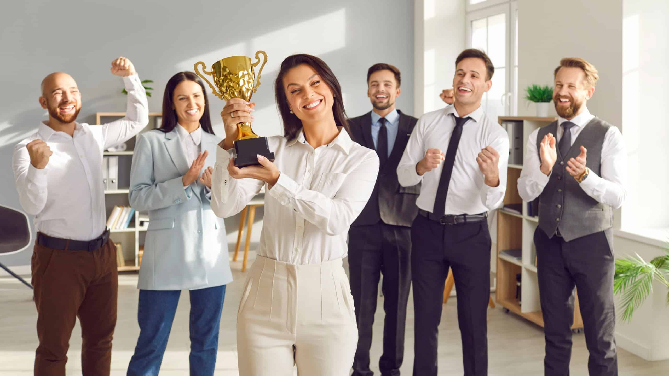 Happy female leader receives business award. Smiling woman winner holding golden trophy, with team of joyful employees in background cheering, celebrating and congratulating her on great work success