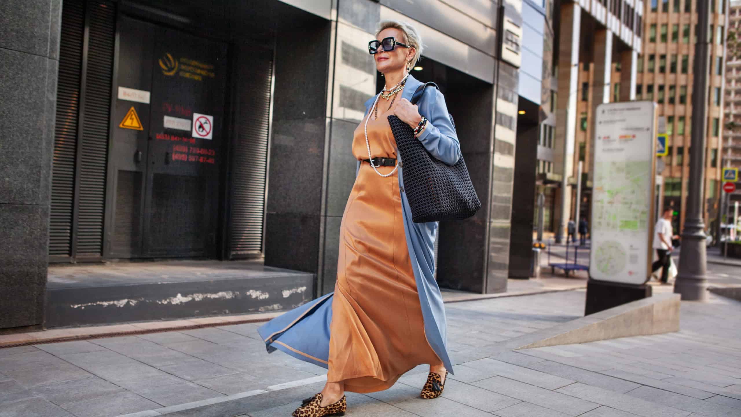 Stylish Mature Woman Walking City Street In Fashionable Clothes, Wearing Long Beige Silk Dress, Blue Cape Coat, Carrying Large Black Handbag, Sunglasses And Trendy Loafers