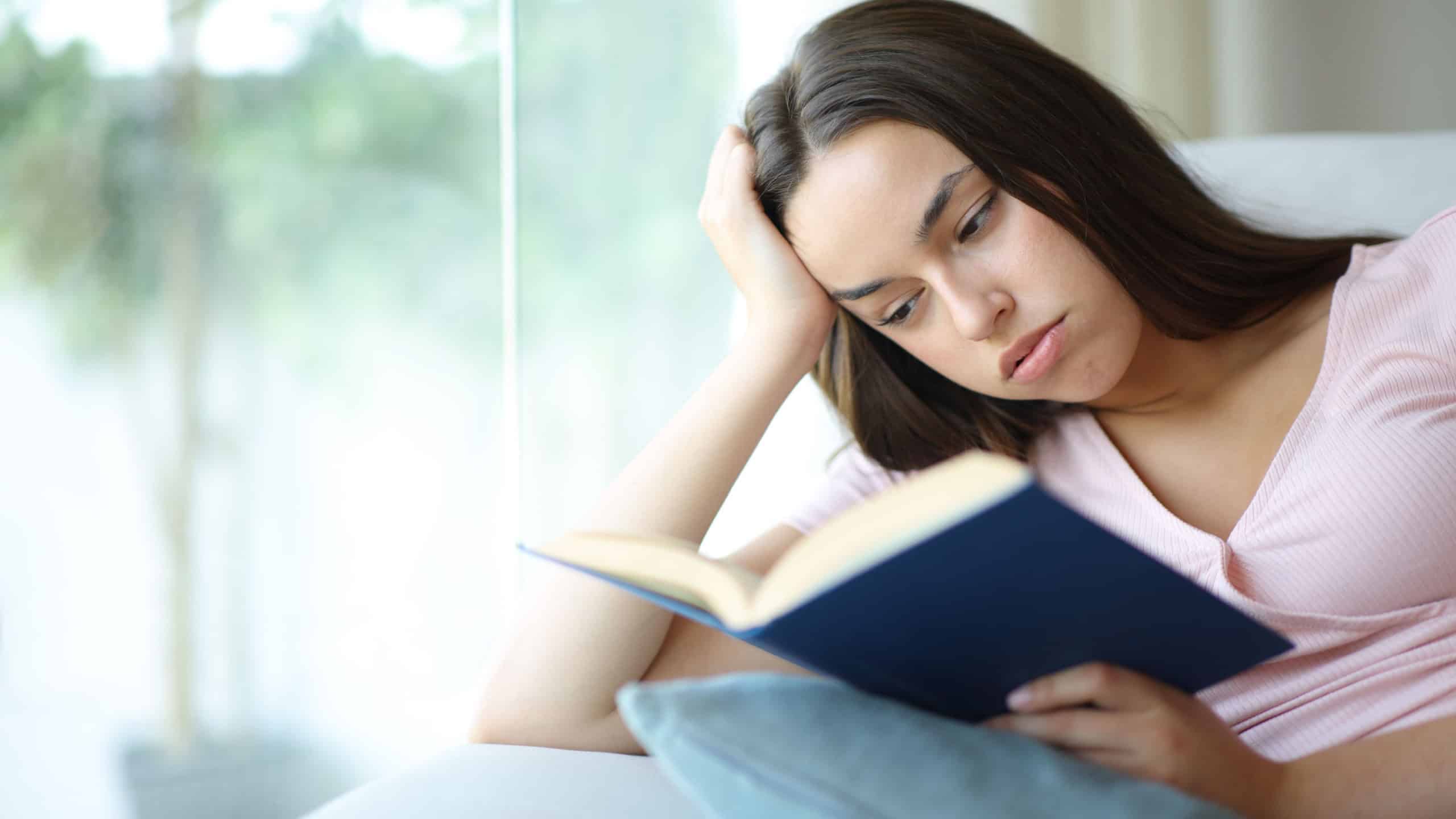 Bored woman is reading a paper book sitting on a couch at home