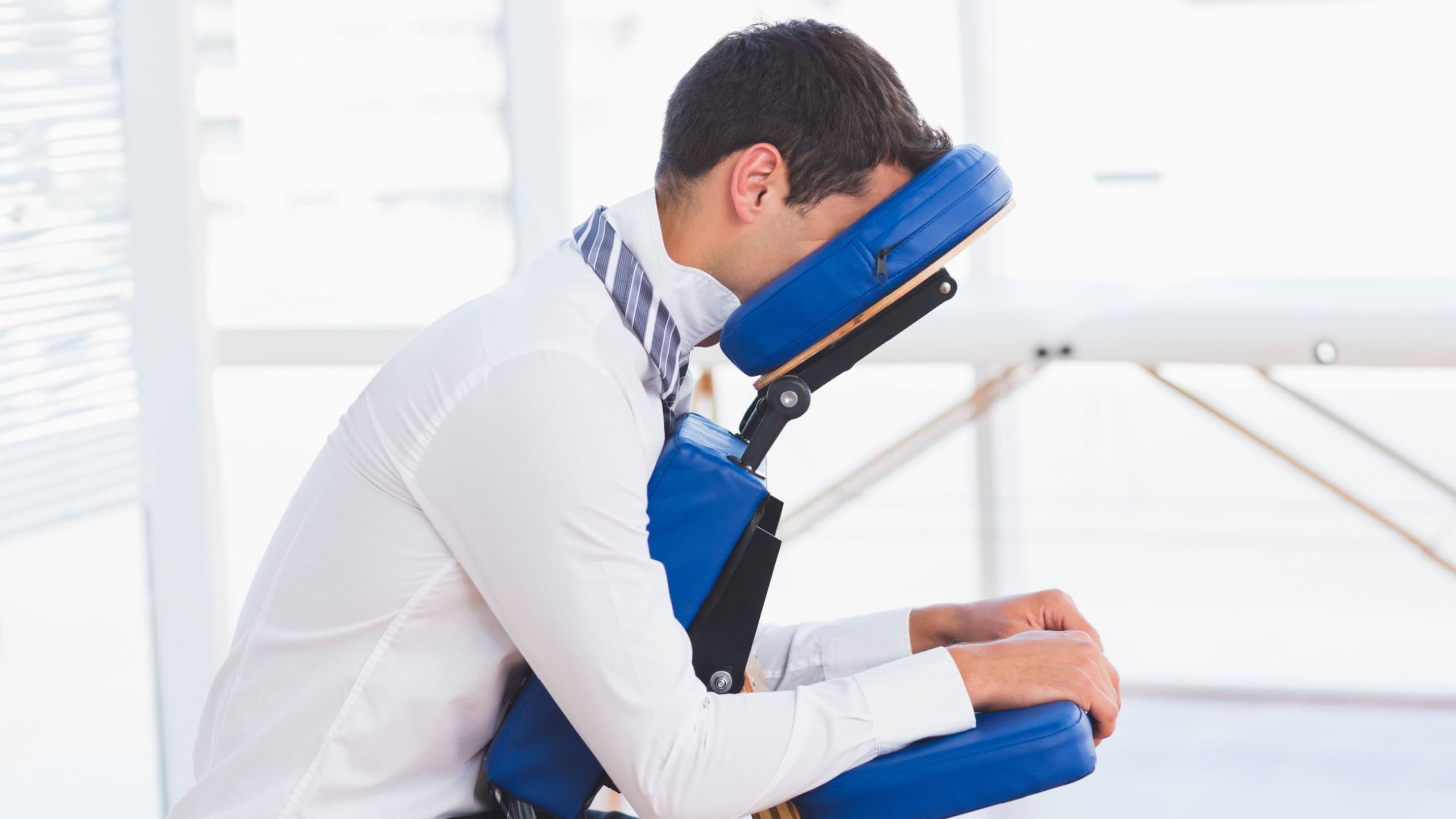 Man in business attire sitting on blue massage chair in wellness room leaning forward. Corporate wellness, relaxation, therapy, comfort, office environment, health, stress relief