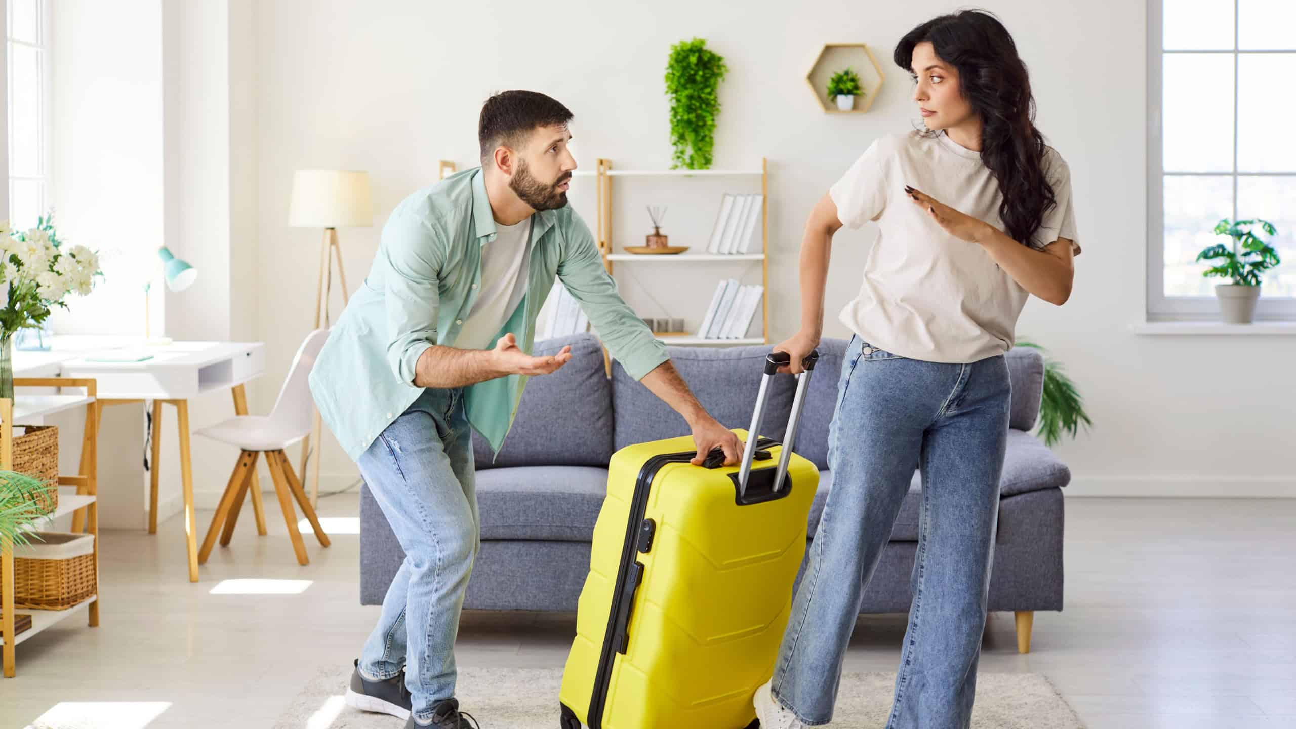 Couple in argument at home. The upset woman with suitcase prepares to leave as the man pleads, showing relationship conflict, stress, and emotions in a modern living room. Theme breakup.