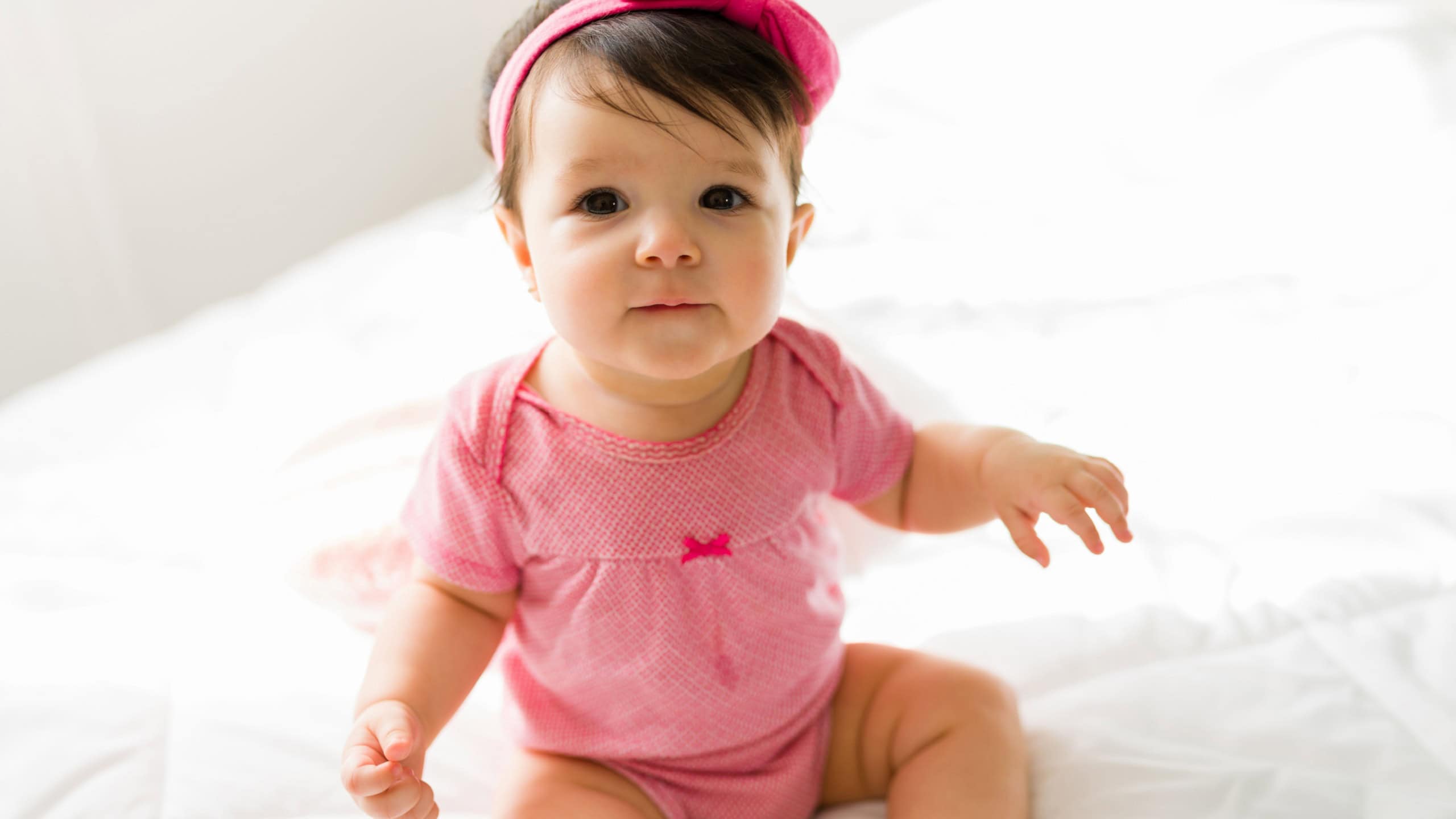 Portrait of a beautiful cute baby girl with a pink headband and onesie sitting on a white bed while looking at the camera