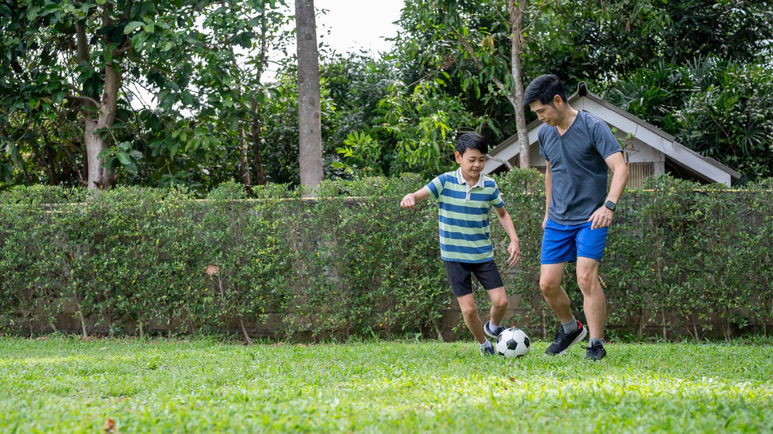 Father and son having fun playing soccer or football in their home's backyard or green grass field. Man and boy, Adult with kid, Sports activity.