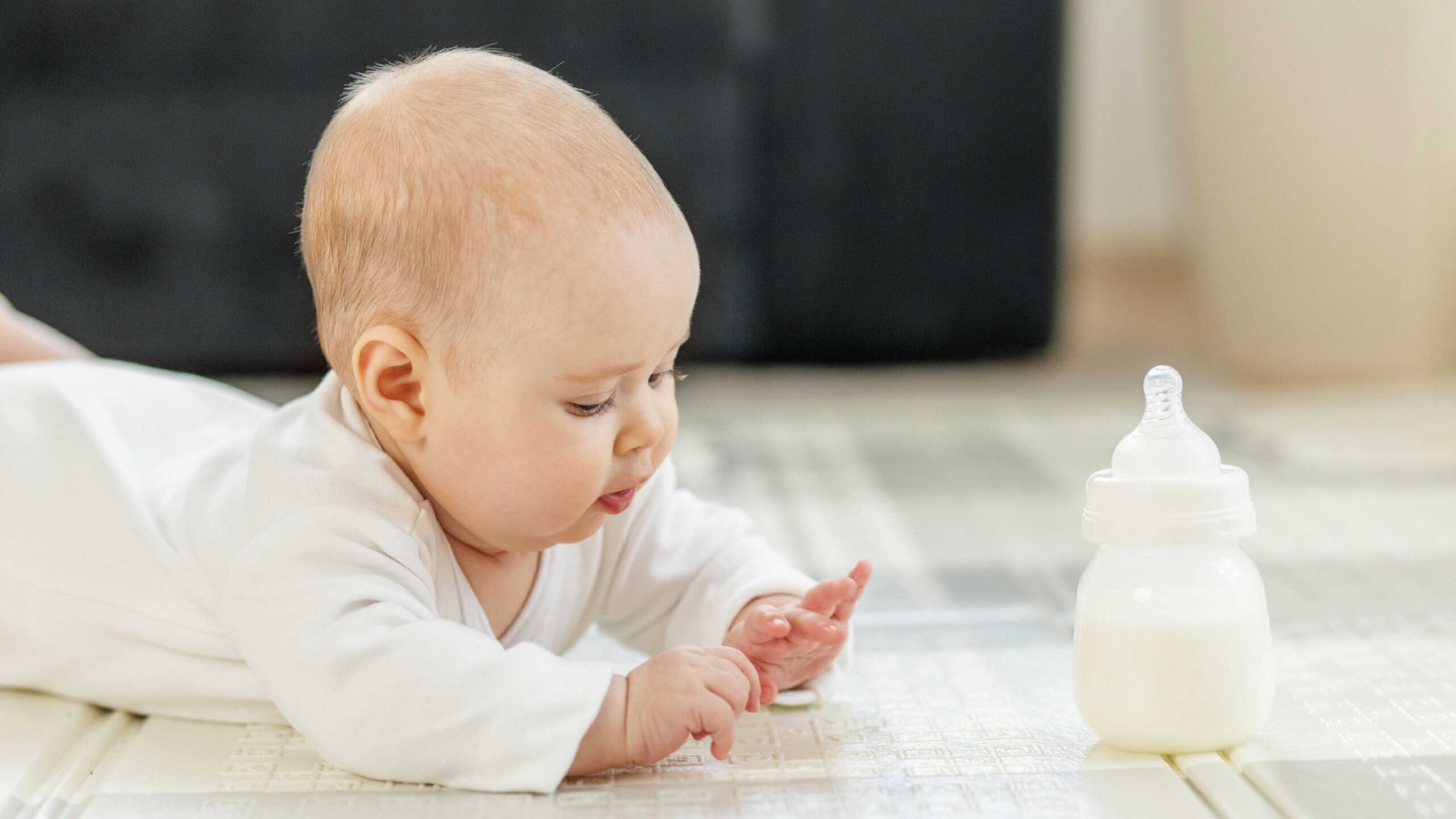 Curious baby in white clothes lying on tummy, exploring surroundings while baby milk bottle stands nearby on clean floor.