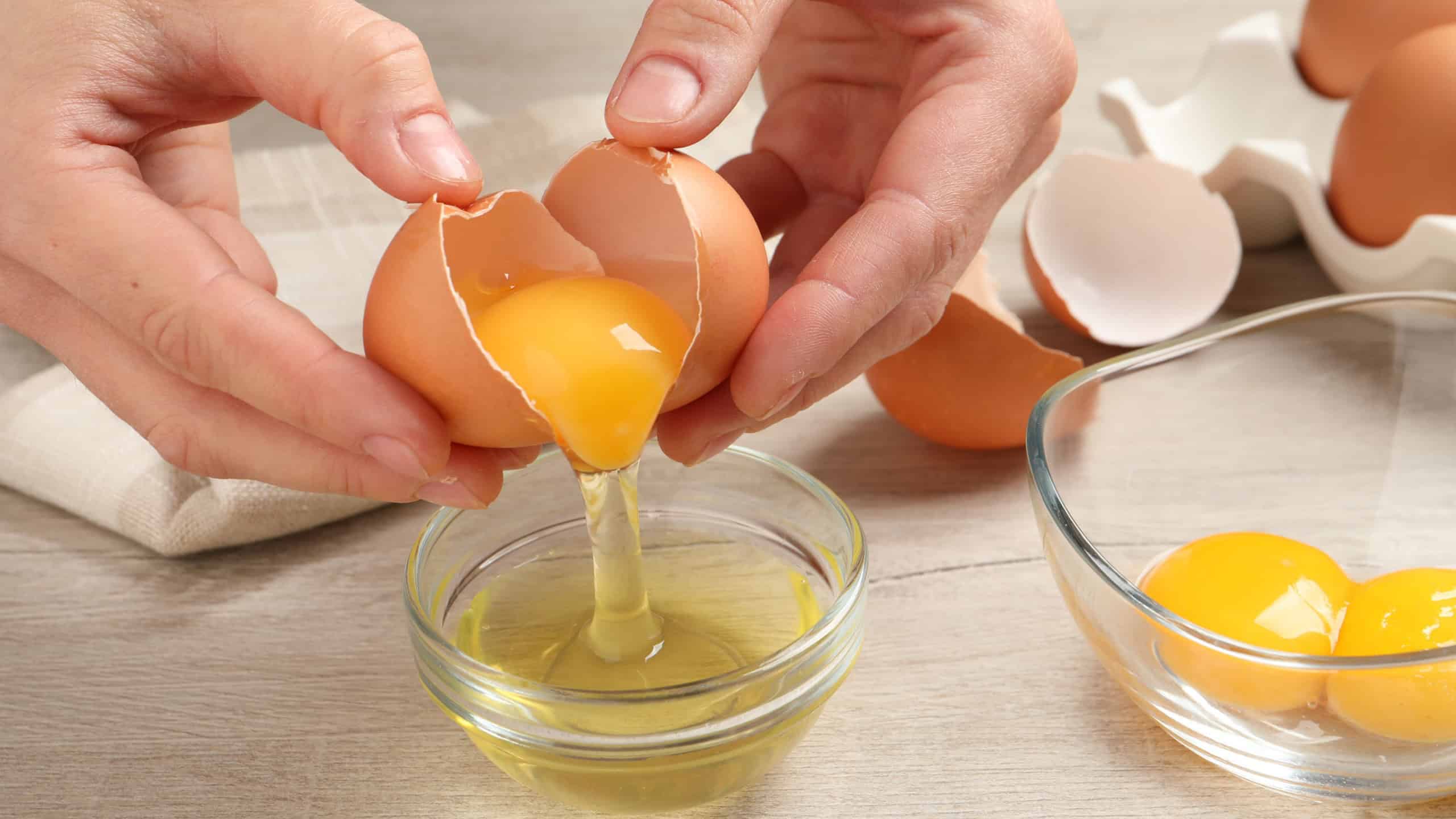 Woman separating egg yolk from white over glass bowl at wooden table, closeup