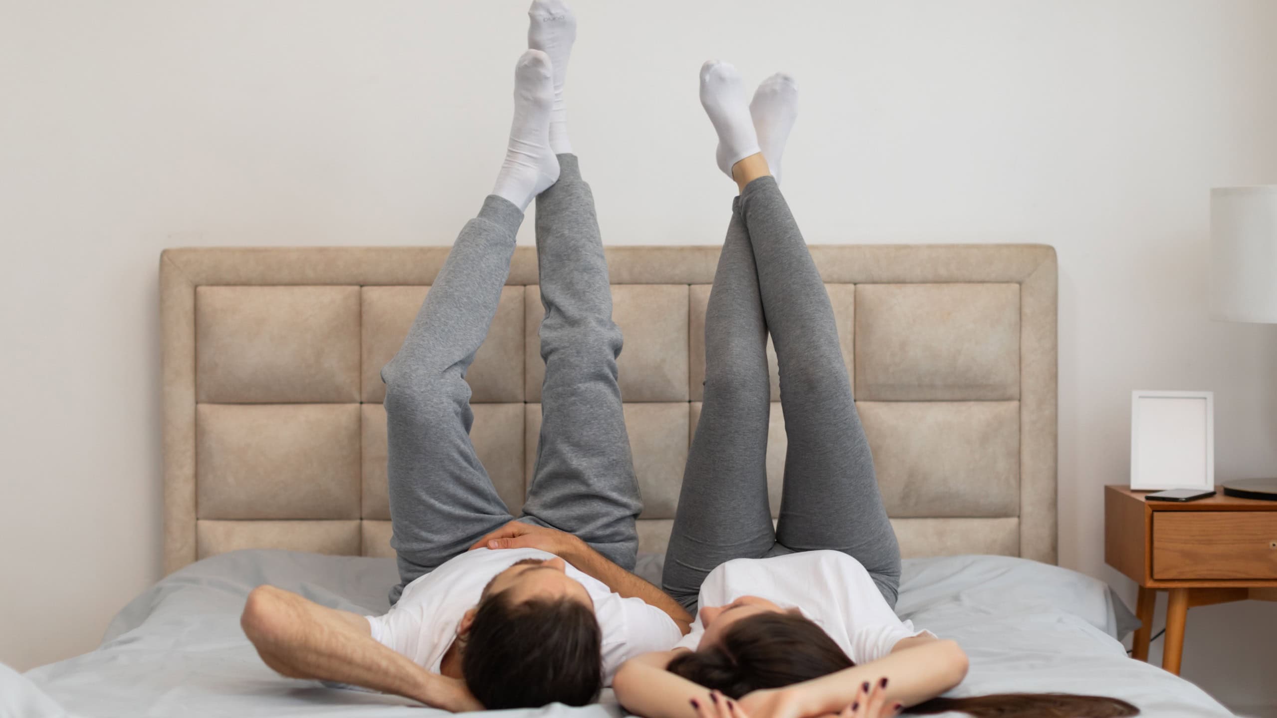 Playful and relaxed couple enjoys light-hearted moment in bed, lying on their backs with their legs up in the air, suggesting comfort and ease