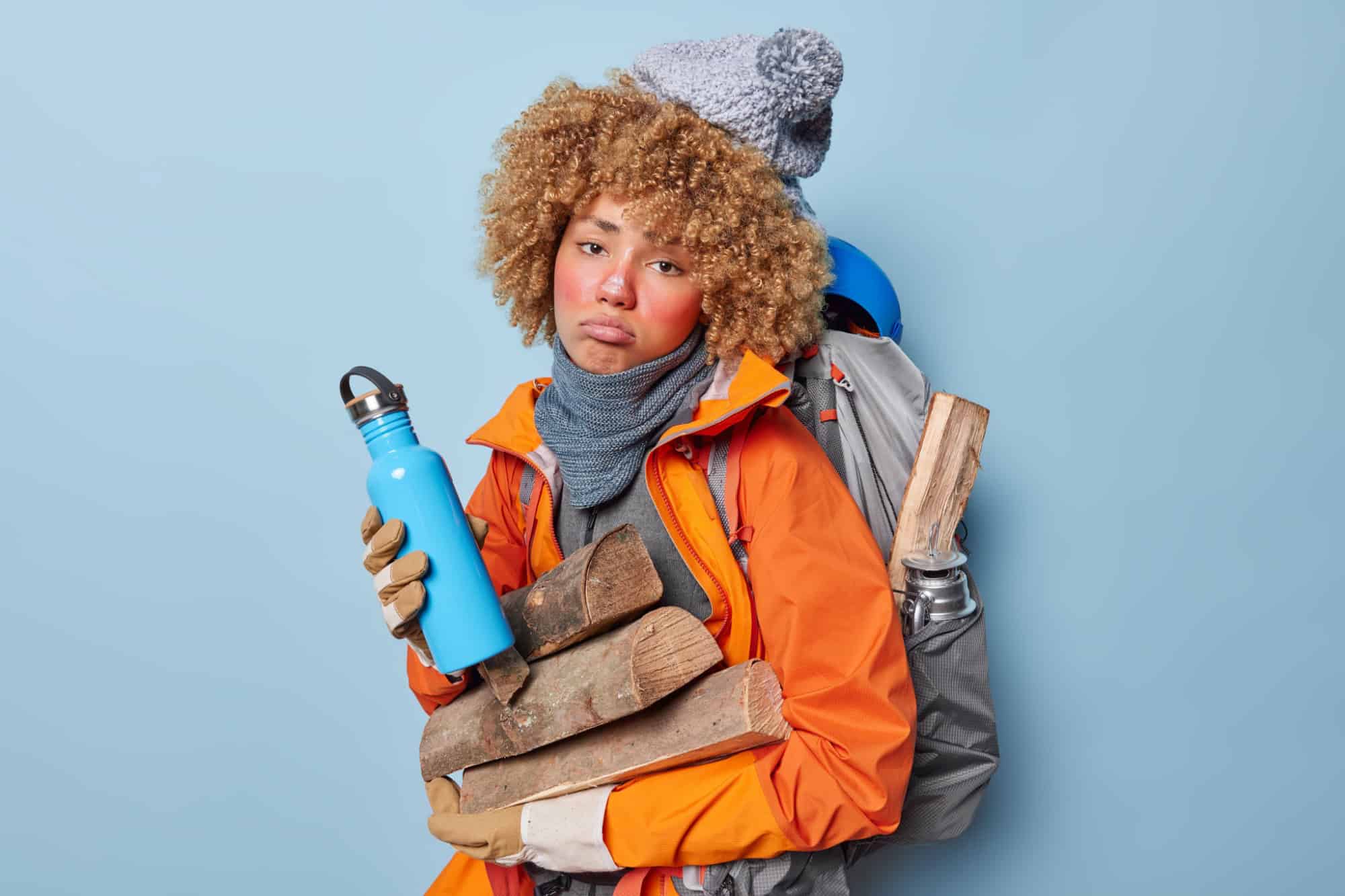 Sad unhappy woman looks discontent holds thermos with hot beverage carries wood for making fire dressed in winter clothes carries rucksack on back feels tired after wandering isolated over blue wall