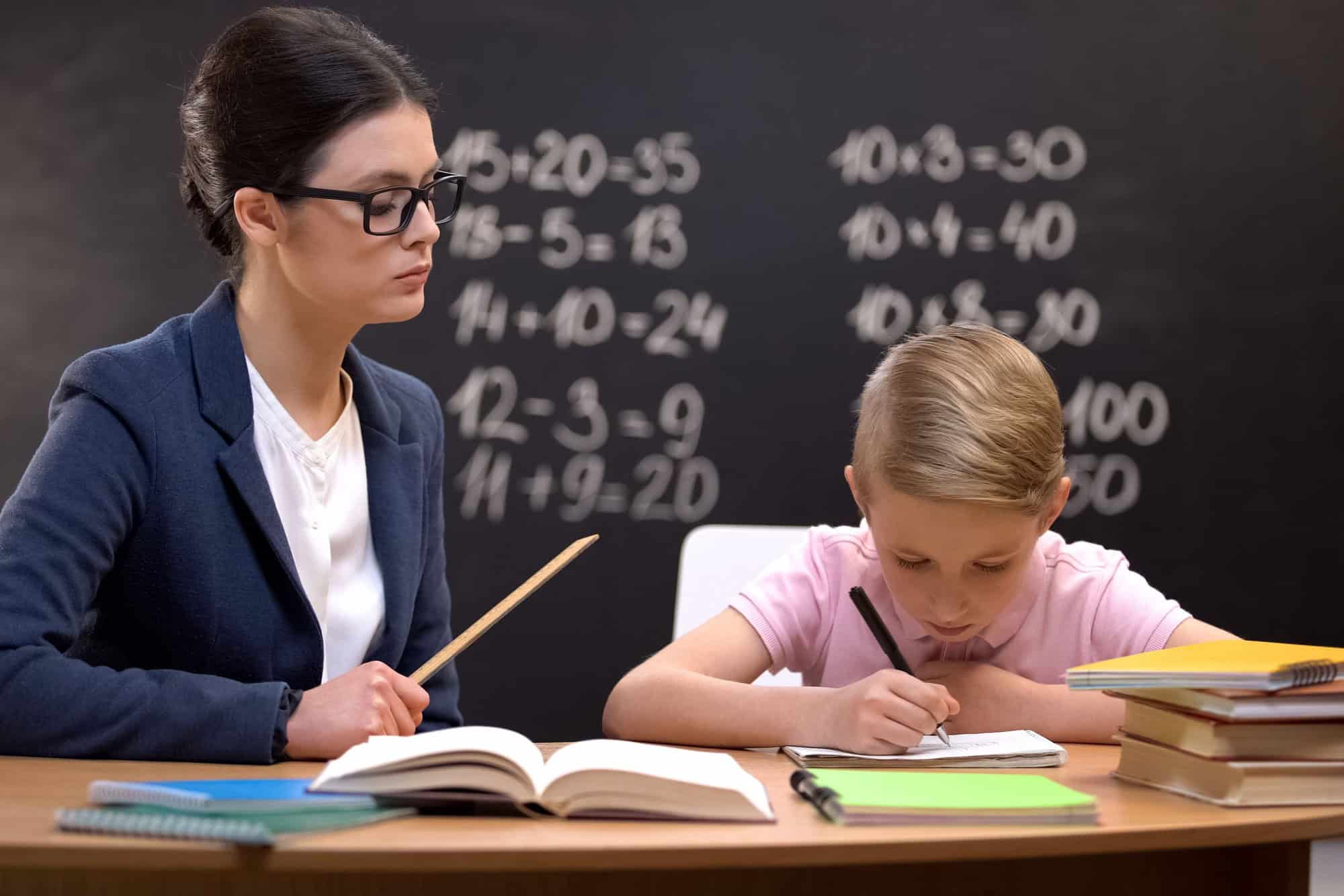 Strict teacher with ruler watching schoolboy doing his homework, control