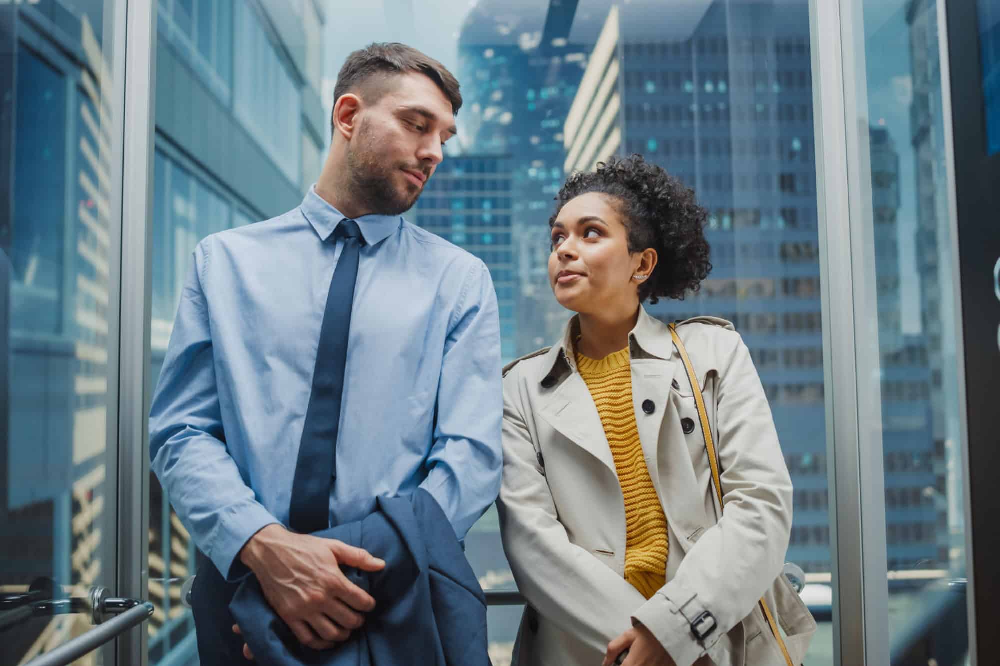 Two Office Colleagues Looking at Each Other While Riding in Glass Elevator in a Modern Business Center. Caucasian Male Specialist and Black Latin American Female Manager Casually Chat in the Lift.