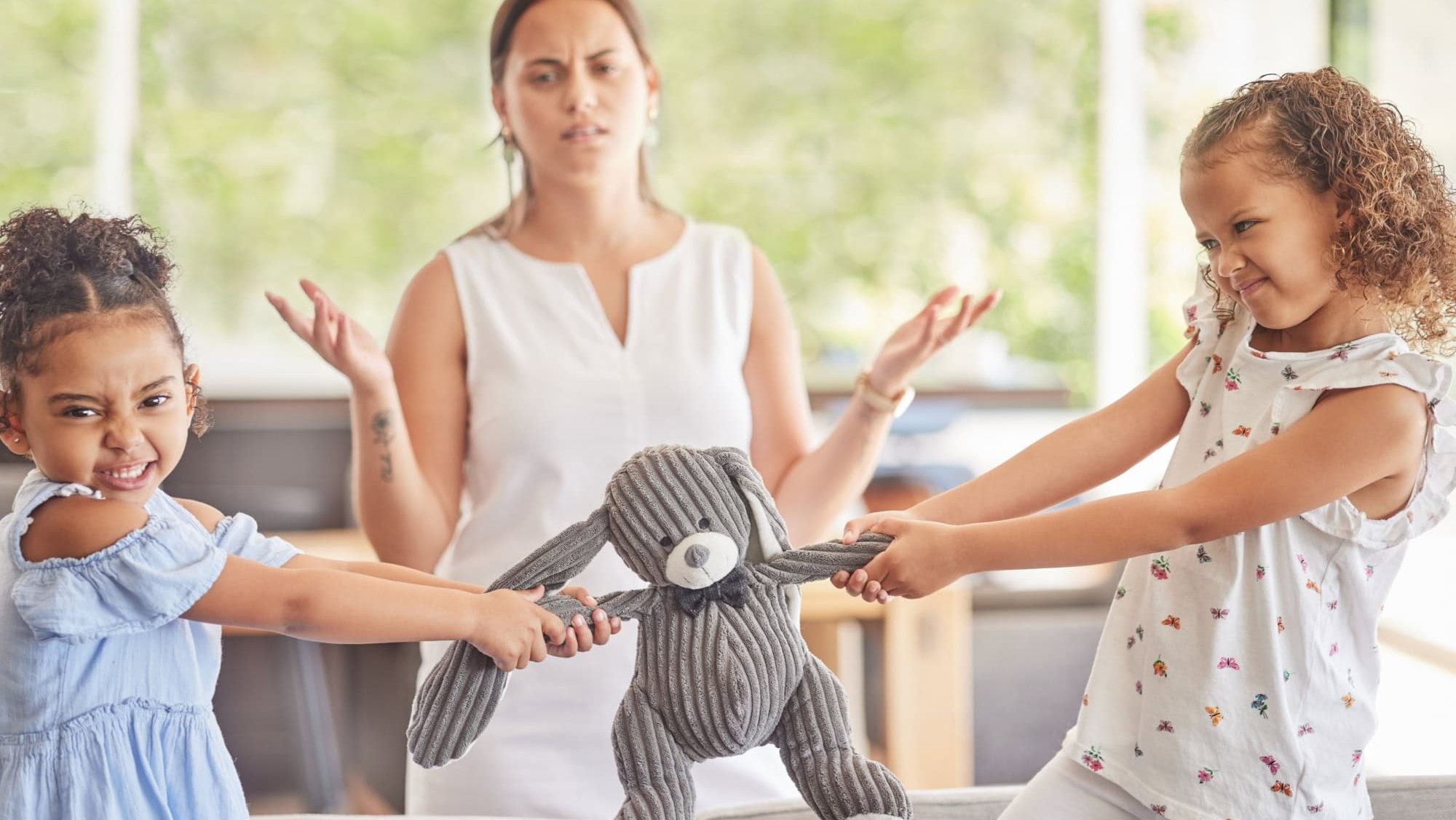 Toys, fight and kids with angry children in conflict while mother confused about family problems or communication. Girl siblings upset about teddy bear with mom trying to talk in home living room