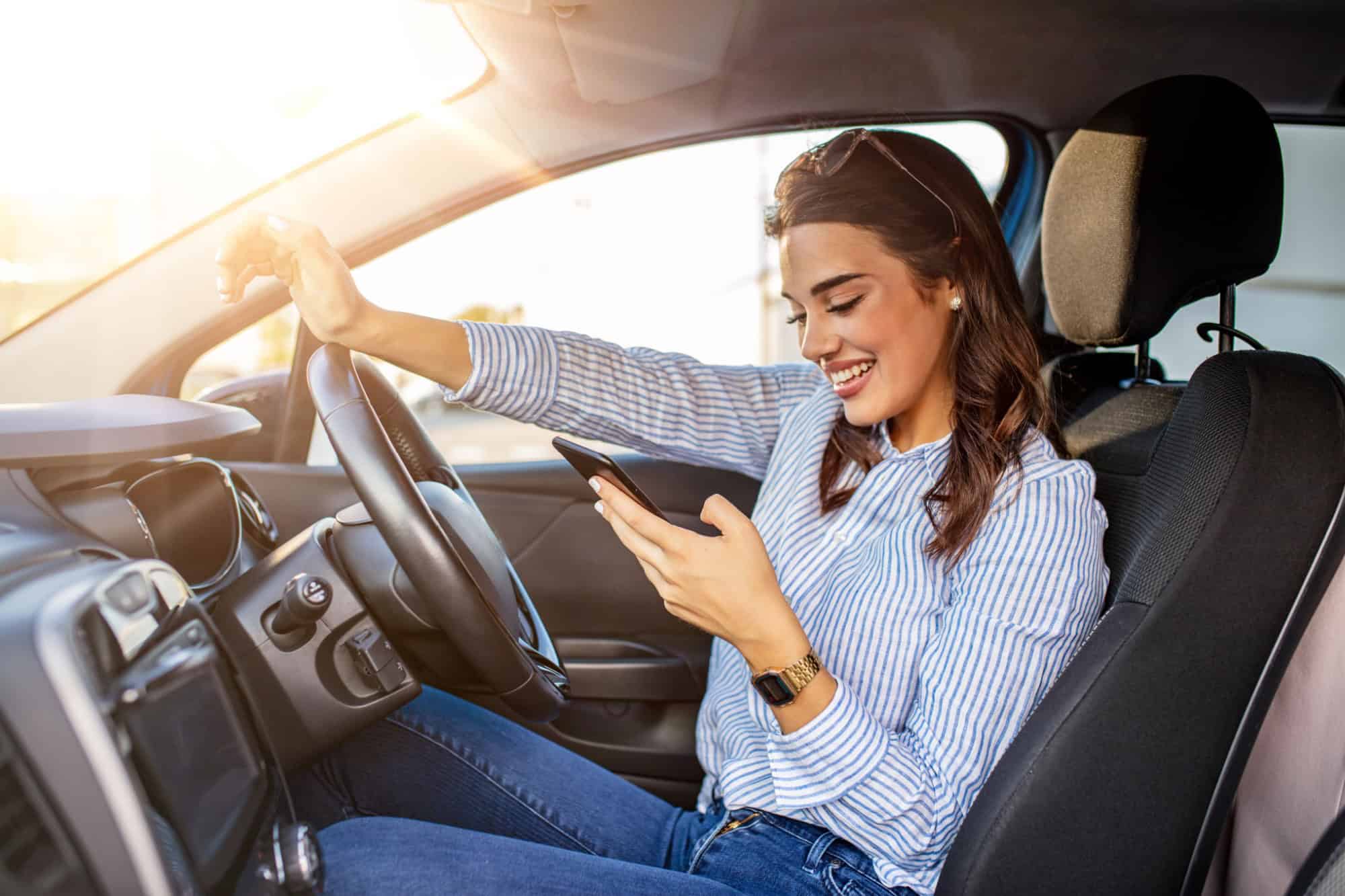Portrait of a young woman texting on her smartphone while driving a car. Business woman sitting in car and using her smartphone. Mockup image with female driver and phone screen