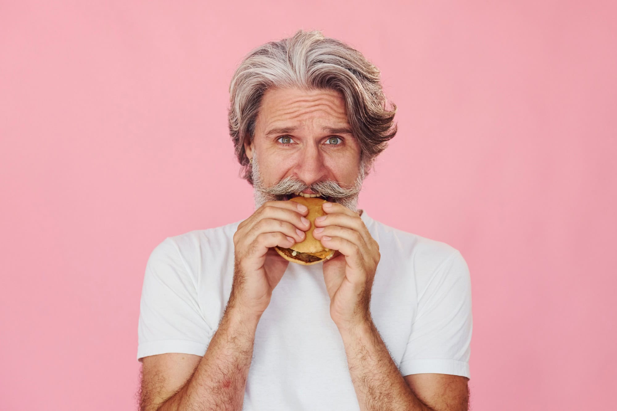 Eats delicious burger. Stylish modern senior man with gray hair and beard is indoors.