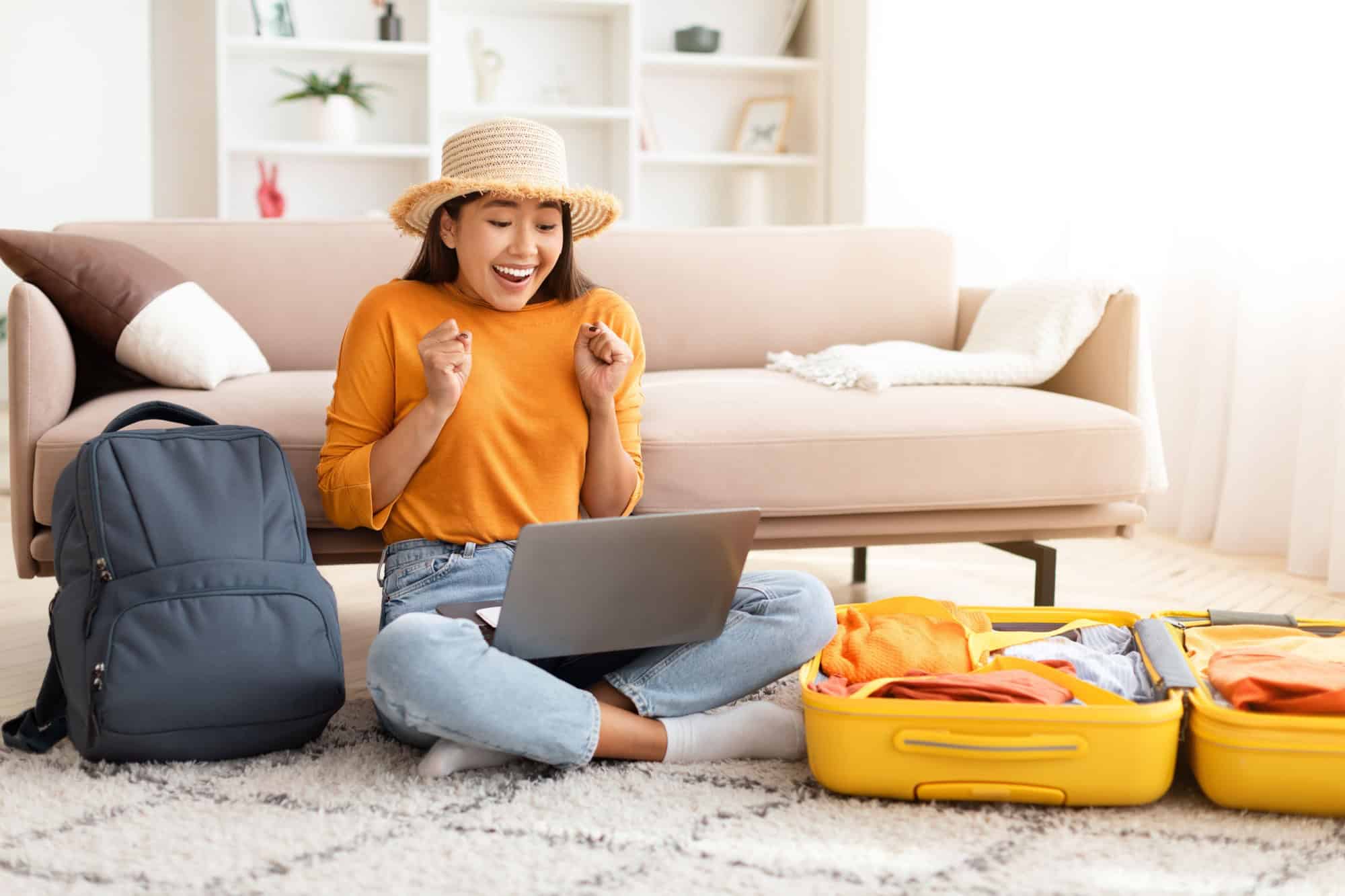 Hot travelling offer, exciting deal. Emotional young asian woman wearing casual clothing and wicker hat traveler sitting on floor, using laptop at home, packing suitcase, backpack, booking tickets