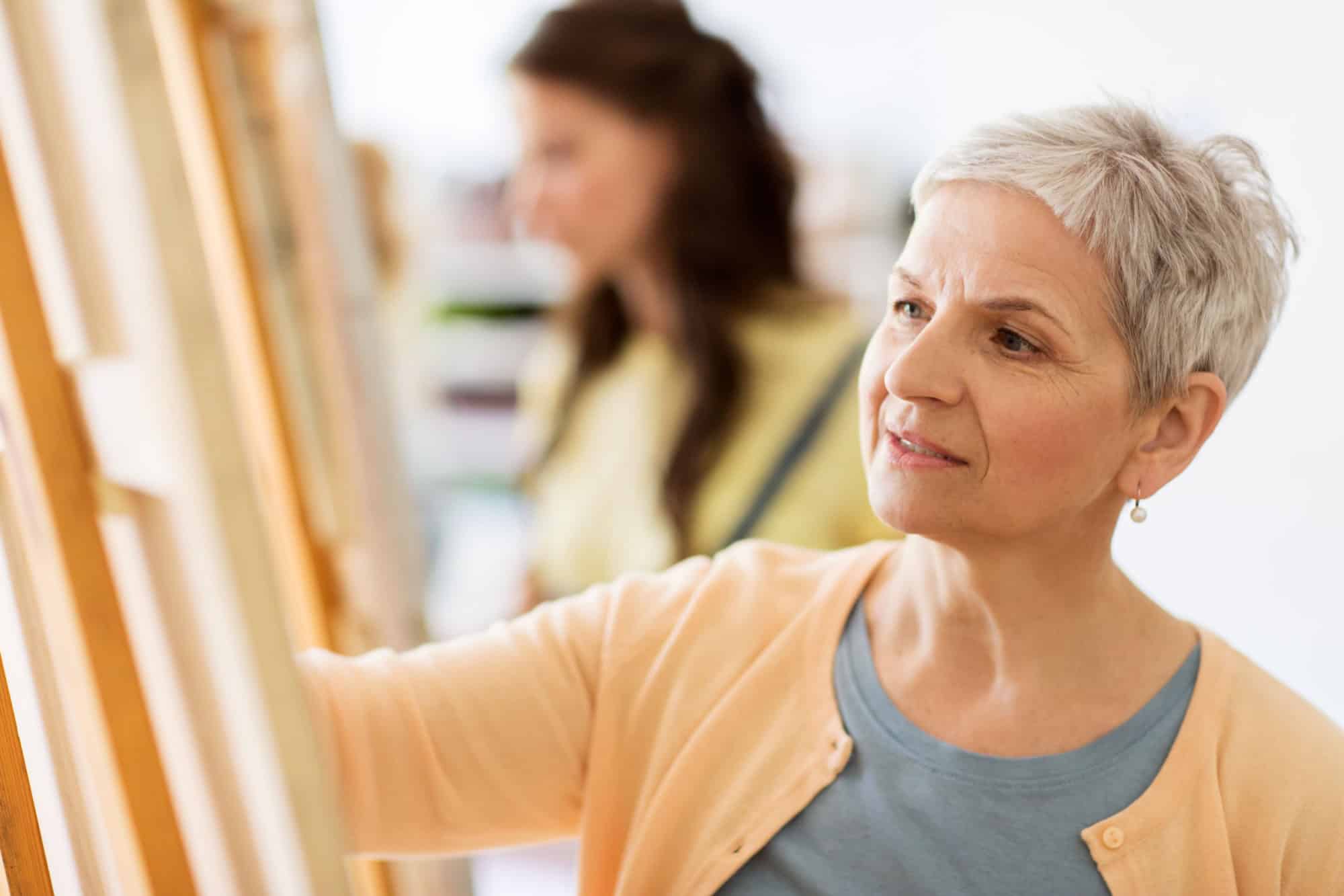 art school, creativity and people concept - happy senior woman artist with easel drawing at studio