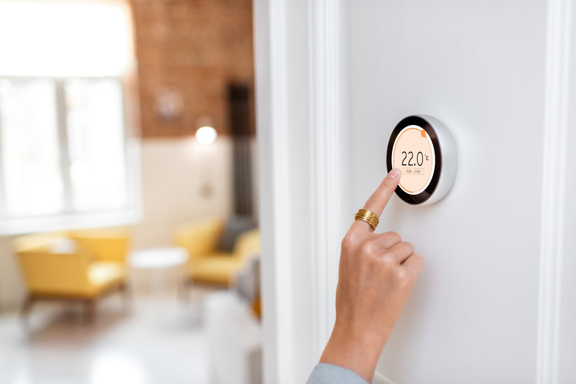 Woman regulating heating temperature with a modern wireless thermostat installed on the white wall at home. Cropped view focused on hand