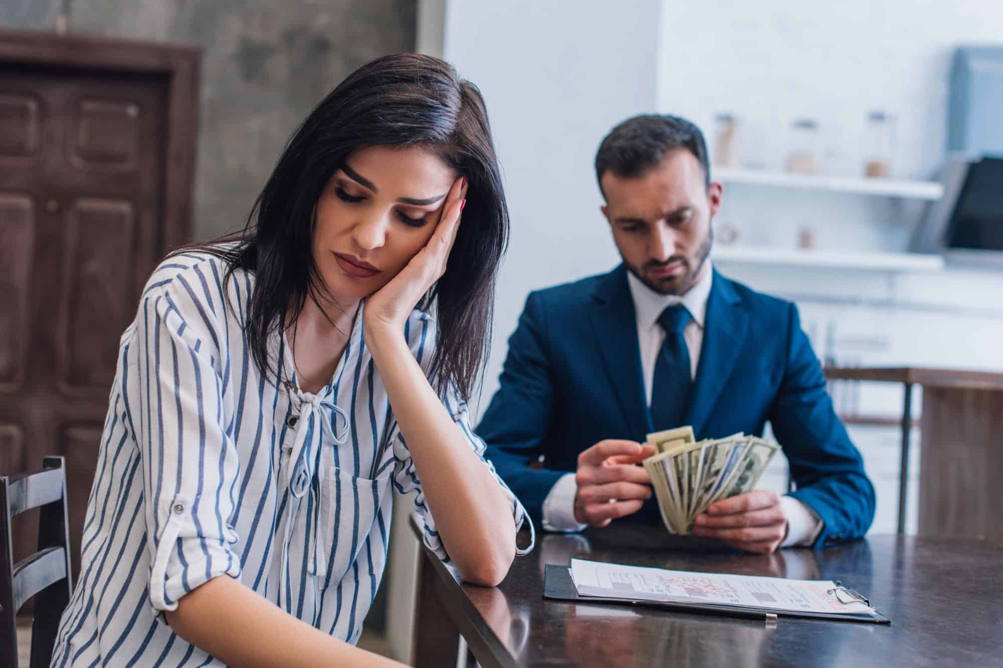 Selective focus of upset woman near documents with collector counting dollar banknotes at table in room