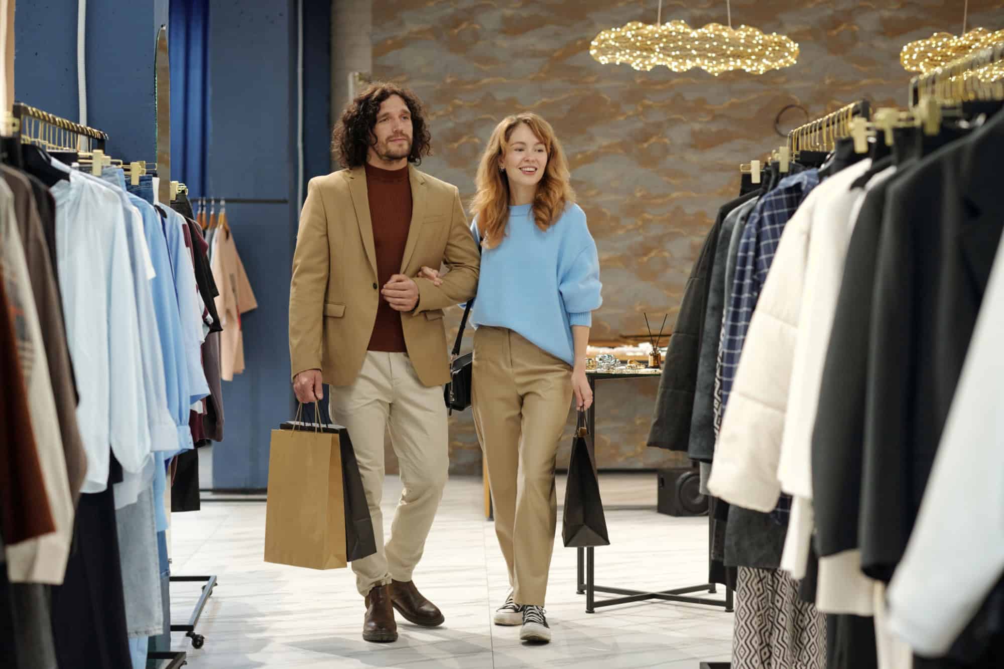 Happy young man and woman in quiet luxury attire moving along racks with new seasonal fashion collection in showroom