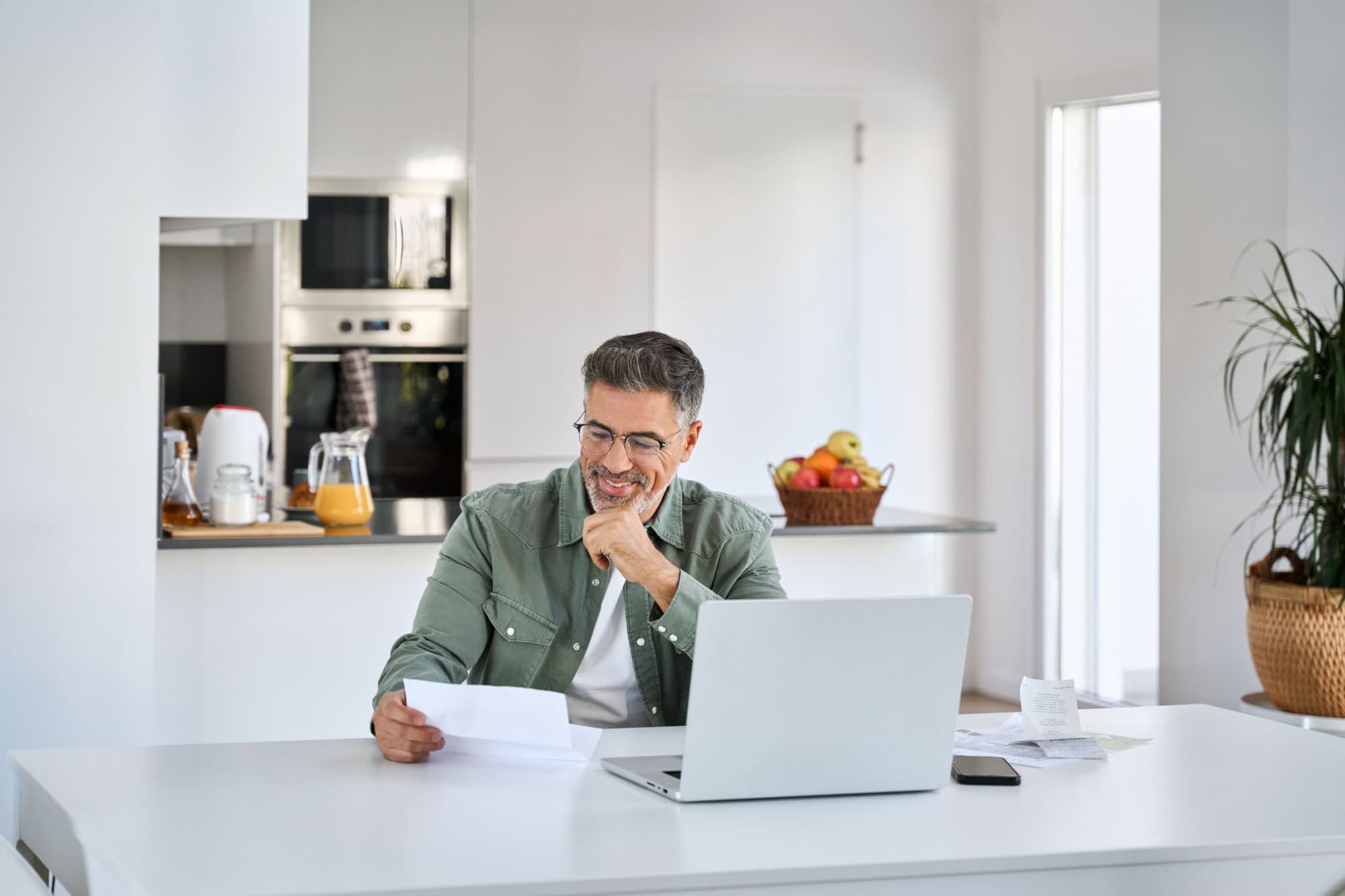 Happy middle aged mature man entrepreneur wearing eyeglasses looking at paper bill checking financial invoice or tax document making online payments working on laptop at home kitchen table.