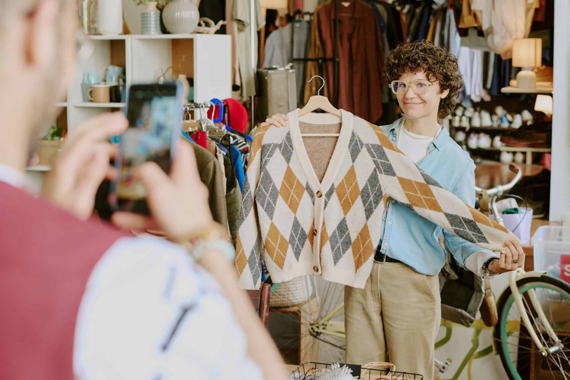 Smiling woman proudly presenting an argyle cardigan in vintage clothing shop while customer capturing the moment with smartphone camera