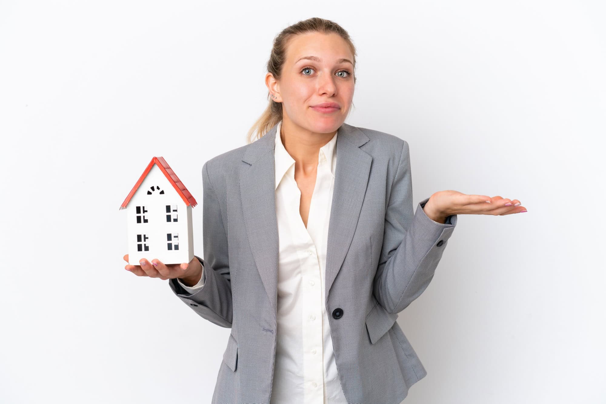 Real estate agent woman holding a toy house isolated on white background having doubts while raising hands