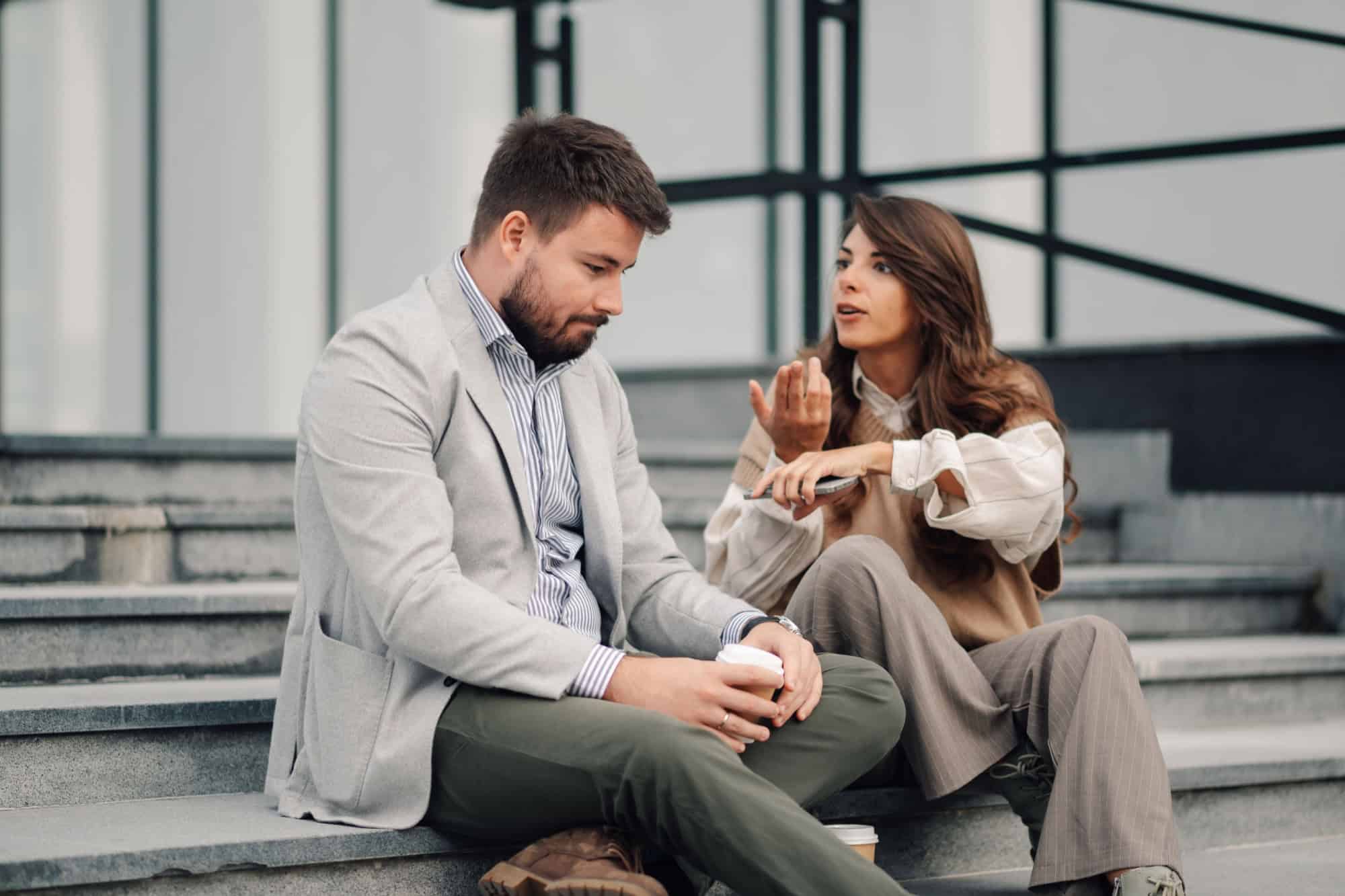 Two colleagues are having a serious conversation about work while sitting on the steps outside, the woman is gesturing while holding her phone and the man is listening intently