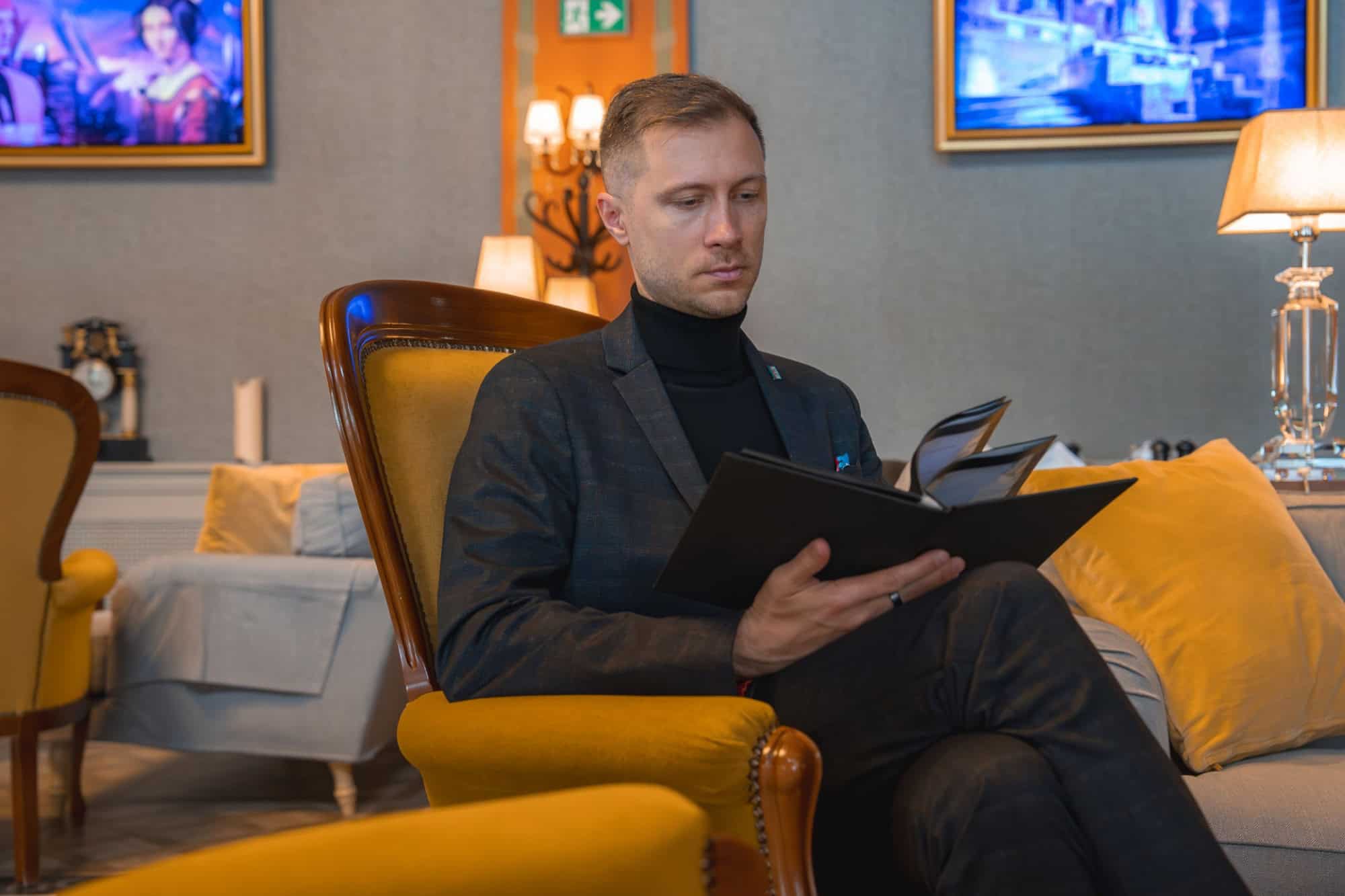 Budapest. Hungary. May 11, 2025. A well dressed man sits in a yellow armchair reading a black book. The room features warm lighting, framed artwork, and sophisticated decor elements.