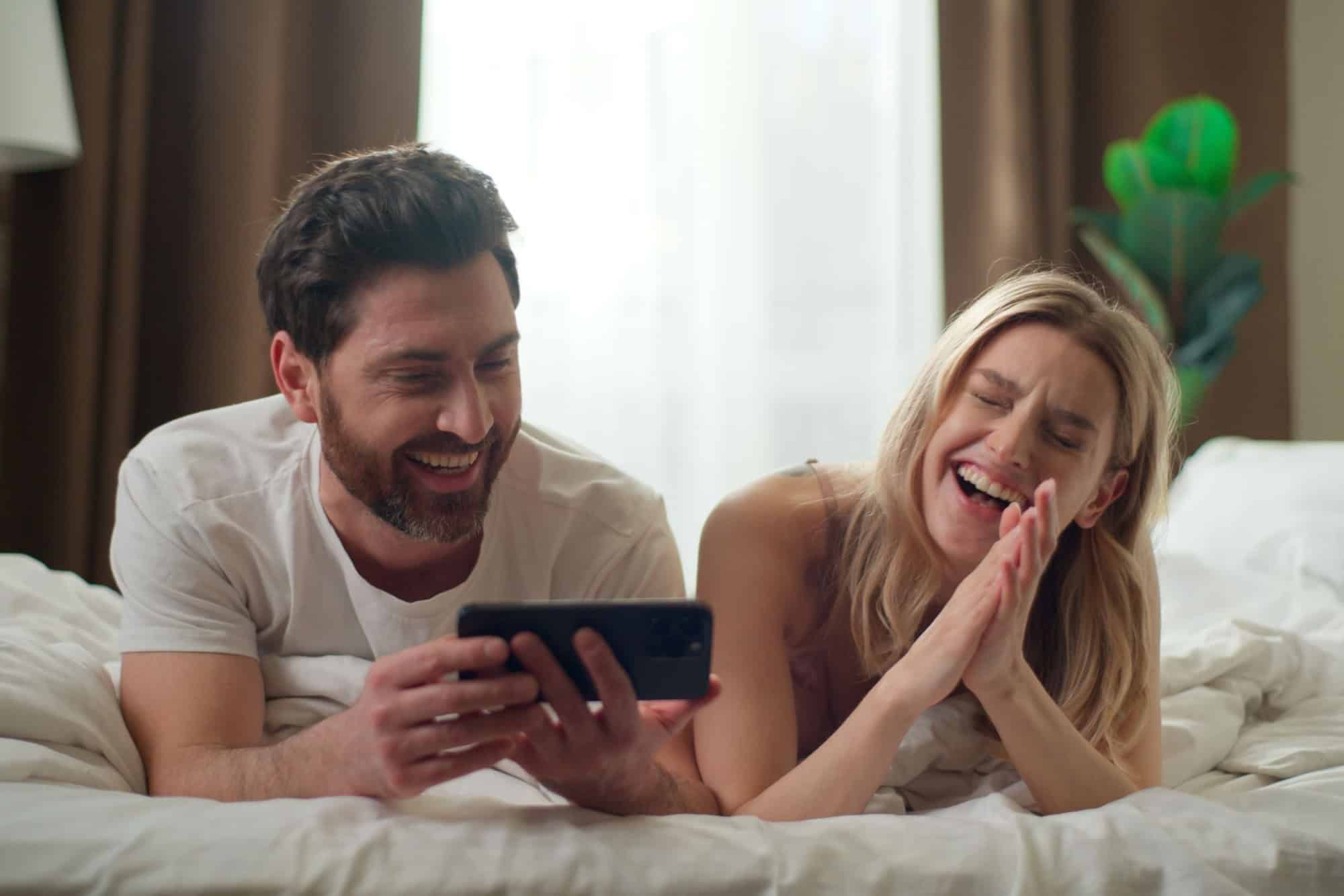 Joyful Caucasian couple family using mobile phone at home girlfriend boyfriend buying online together network positive man scrolling web Internet cyberspace app woman laughing talking bed love morning