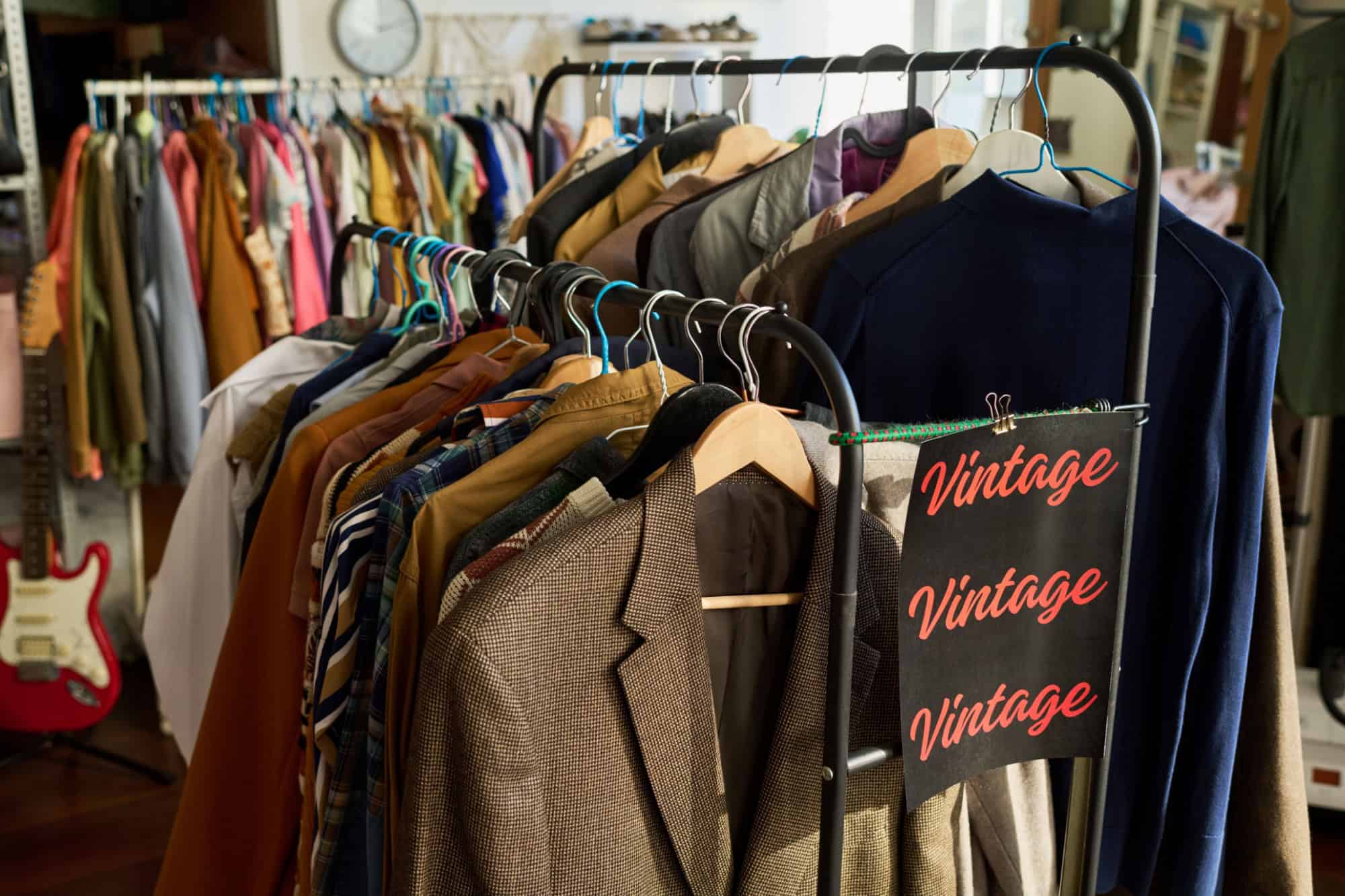 Clothing rack displaying assorted vintage jackets and shirts in thrift shop, garments hanging on wooden and plastic hangers with sign reading vintage, background showing more clothing racks
