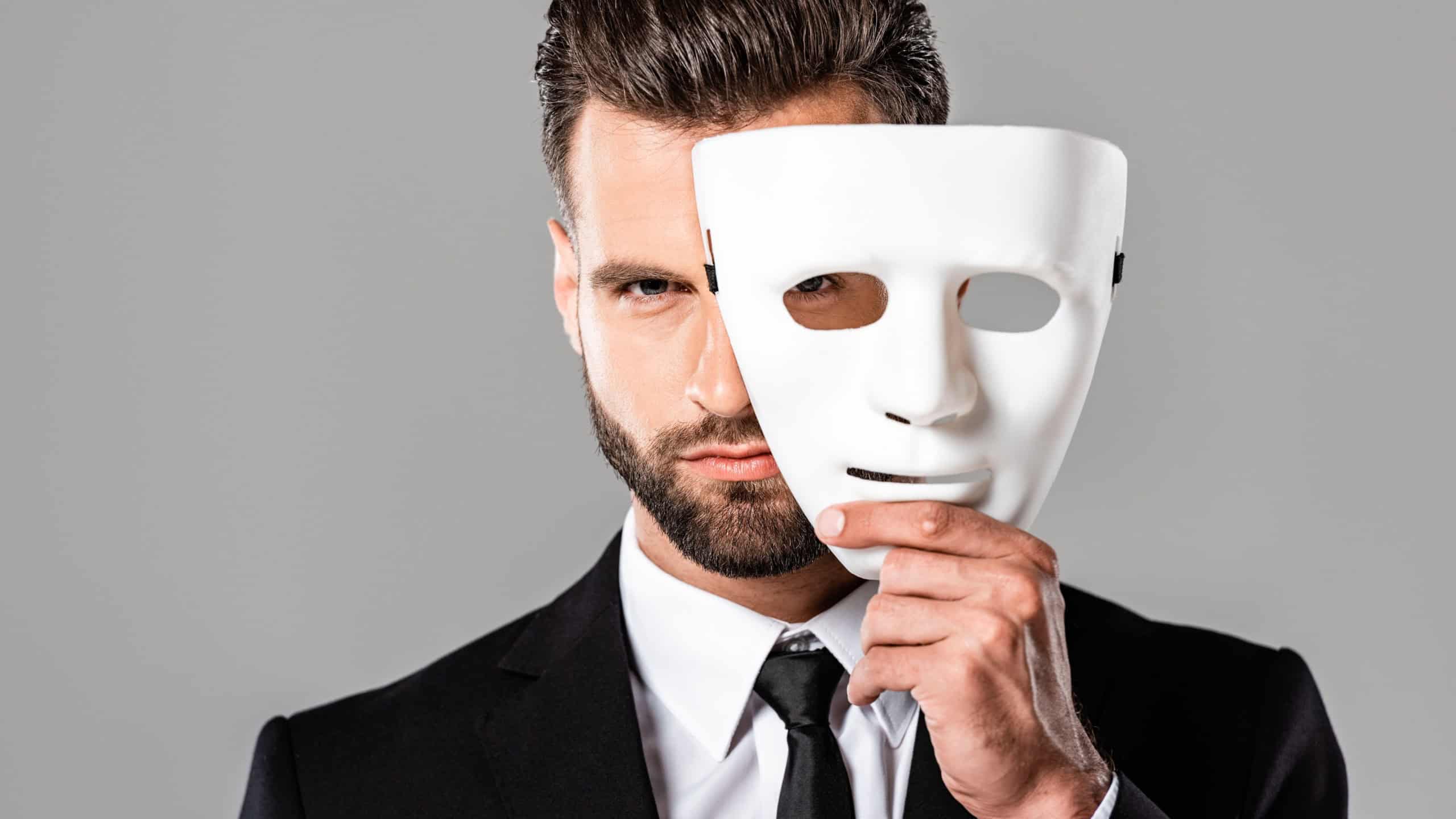 serious handsome businessman in black suit taking off white mask isolated on grey