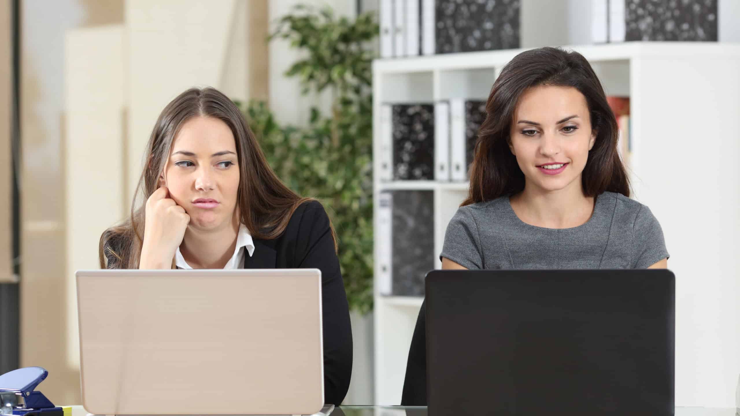 Front view of an envious employee looking her colleague working beside