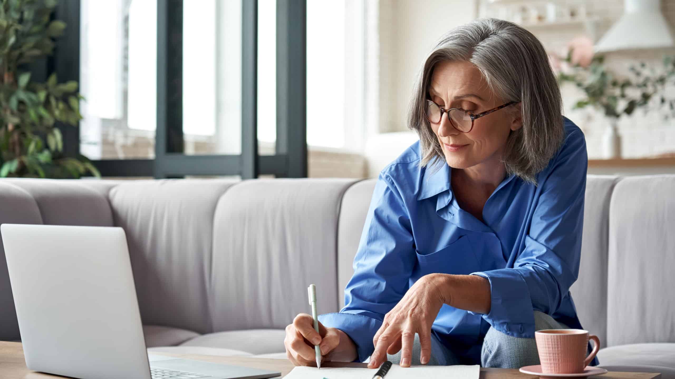 Happy stylish mature old woman remote working from home distance office on laptop taking notes. Smiling 60s middle aged business lady using computer watching webinar sit on couch writing in notebook.