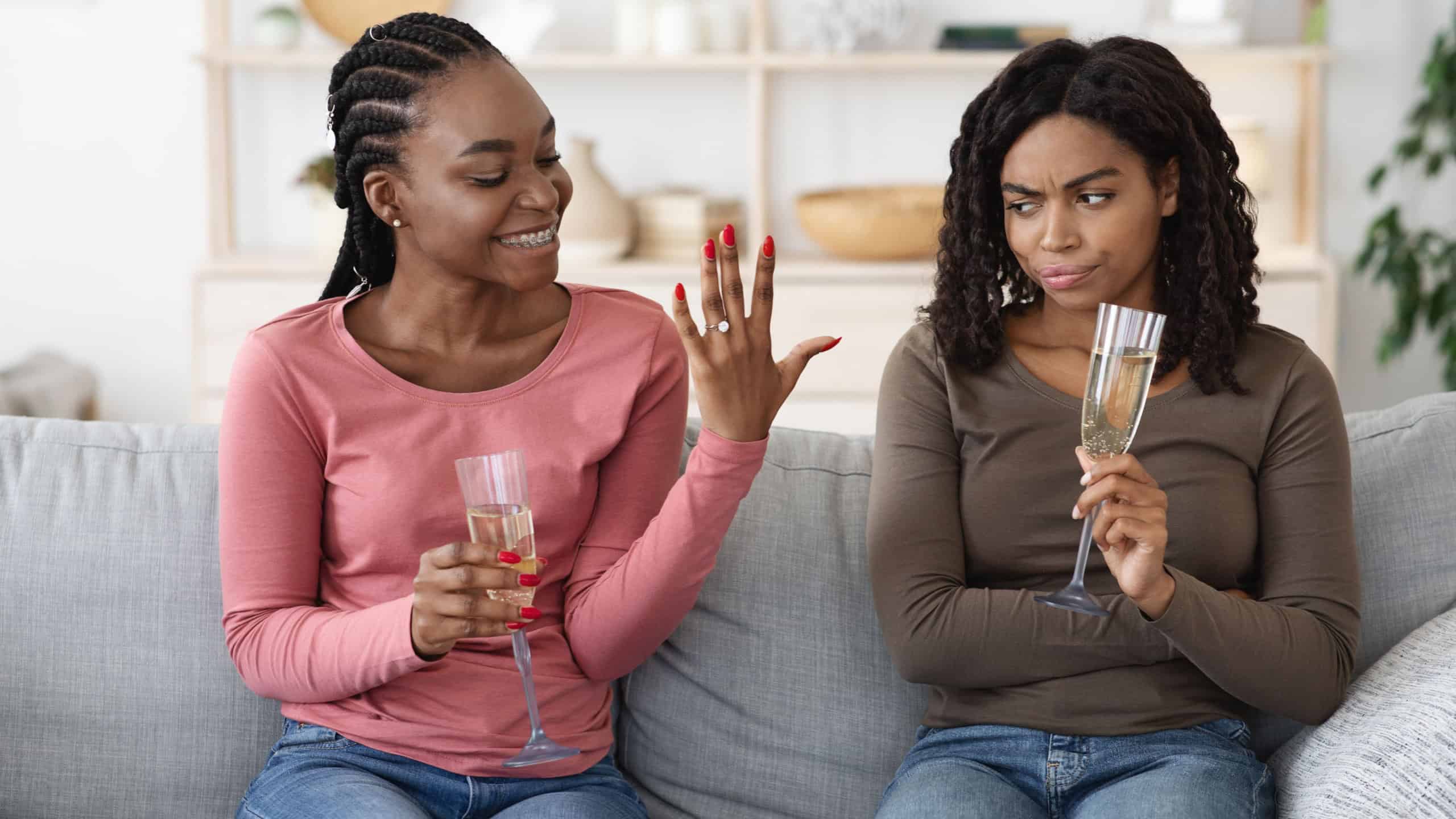 Happy black woman showing her upset and jealous girlfriend engagement ring. Annoyed african american lady cannot share her female friend happiness, drinking champagne together at home