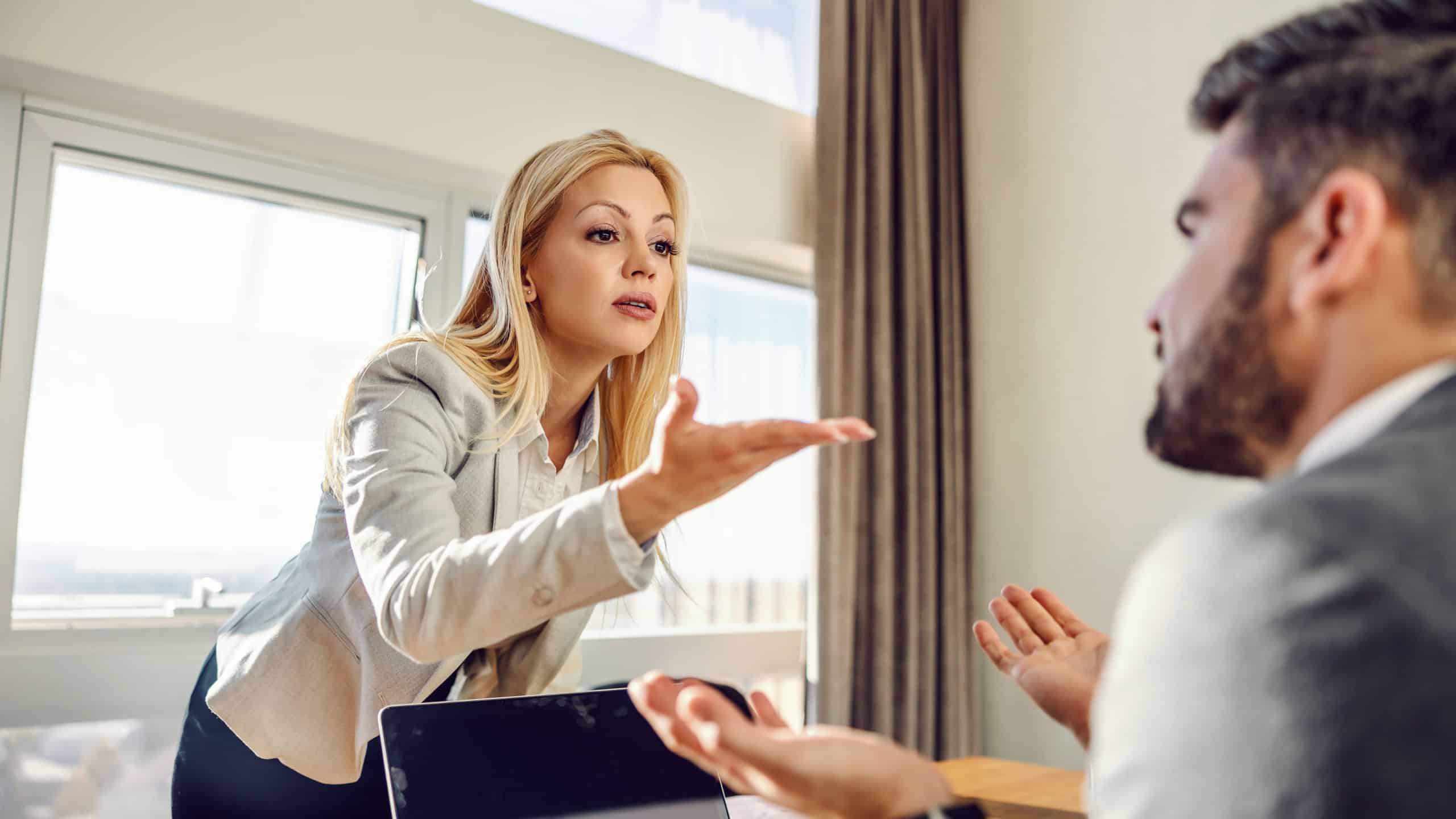 Businesswoman standing at the office and having a disagreement with her colleague who is sitting in elegant clothes in front of a laptop. Issues at work, disagreement