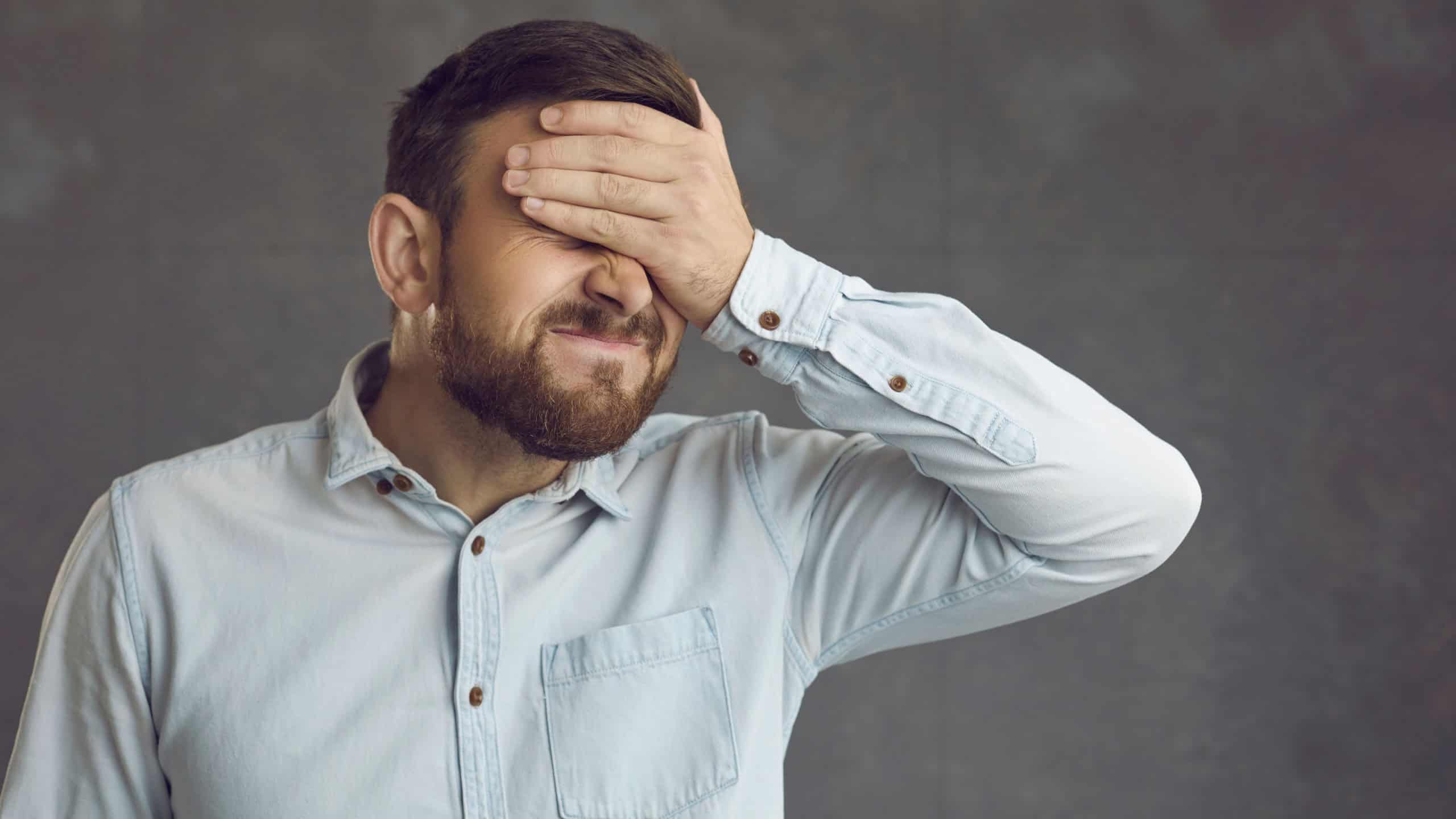 Businessman covers face with hand. Guy facepalms feeling ashamed of terrible mistake or poor memory. Annoyed guilty cringing business manager employee closes face with palm isolated on grey background