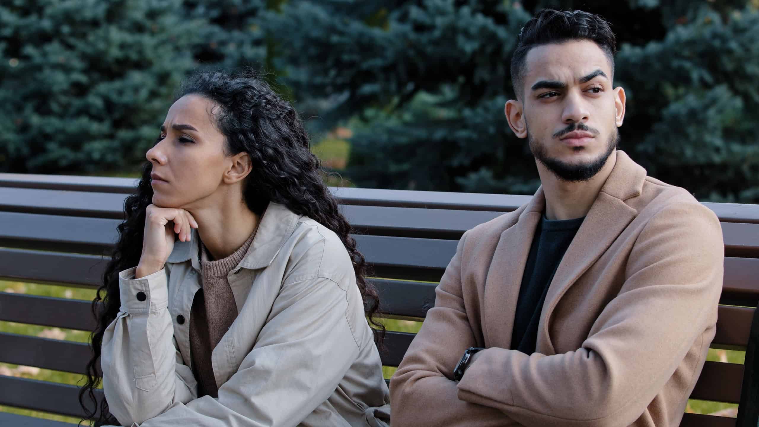 Offended young hispanic couple looking different direction sitting on bench in autumn park annoyed silent guy indignant girl heavily sigh thinking breakup with boyfriend woman in quarrel with beloved