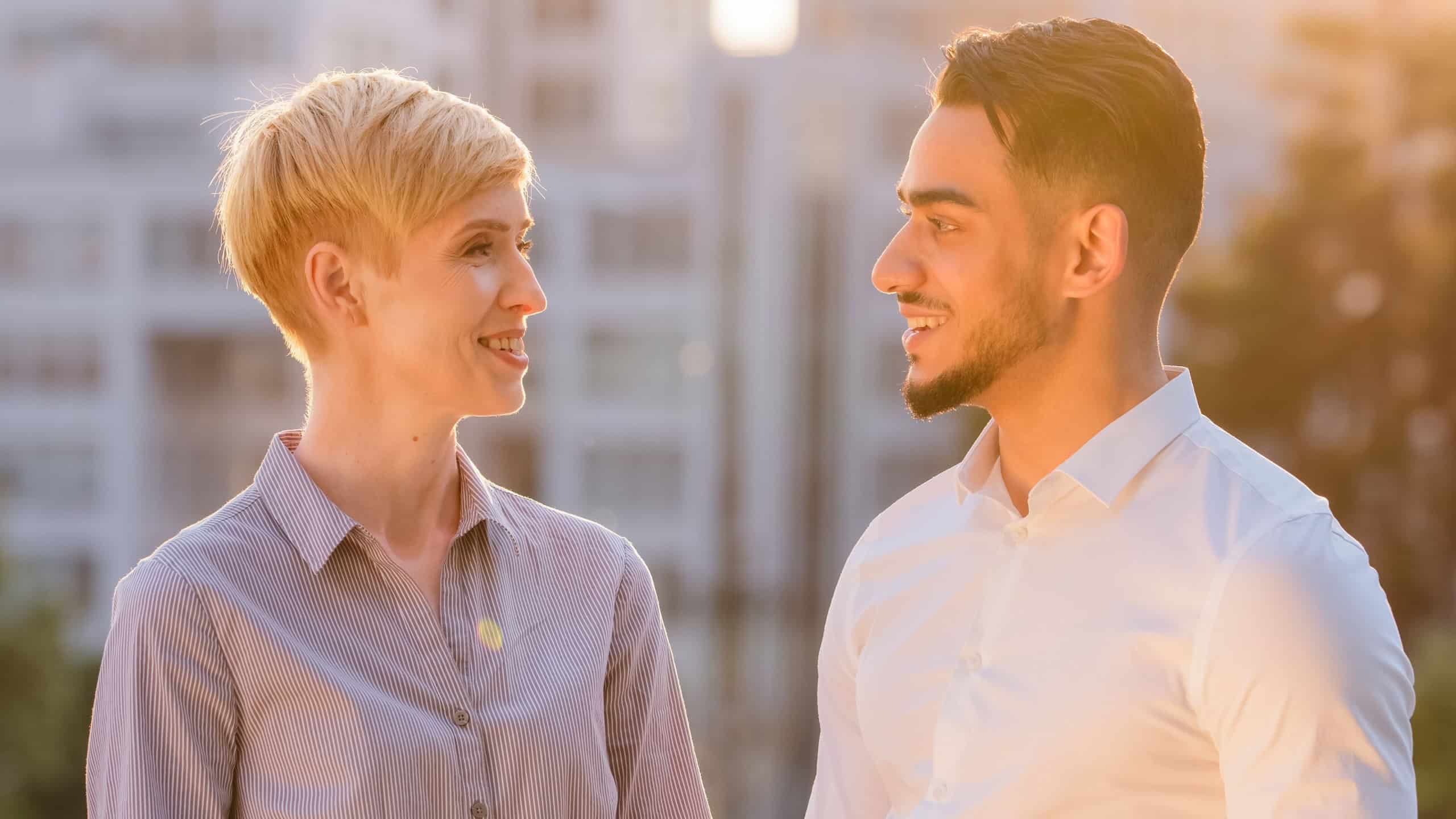 Multiracial business colleague couple arabic hispanic man and mature caucasian woman standing outdoors in sunlight talking conversation negotiation looking at camera smiling nodding heads yes positive